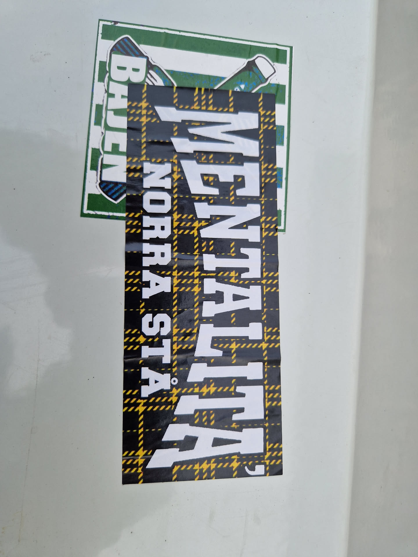 A black and yellow plaid sticker with the word "MENTALITA" written in white bold letters. There is a smaller sticker above it, partially visible, showing green and white colors.  The main sticker has a slightly worn look, suggesting it may have been applied outdoors for a while.