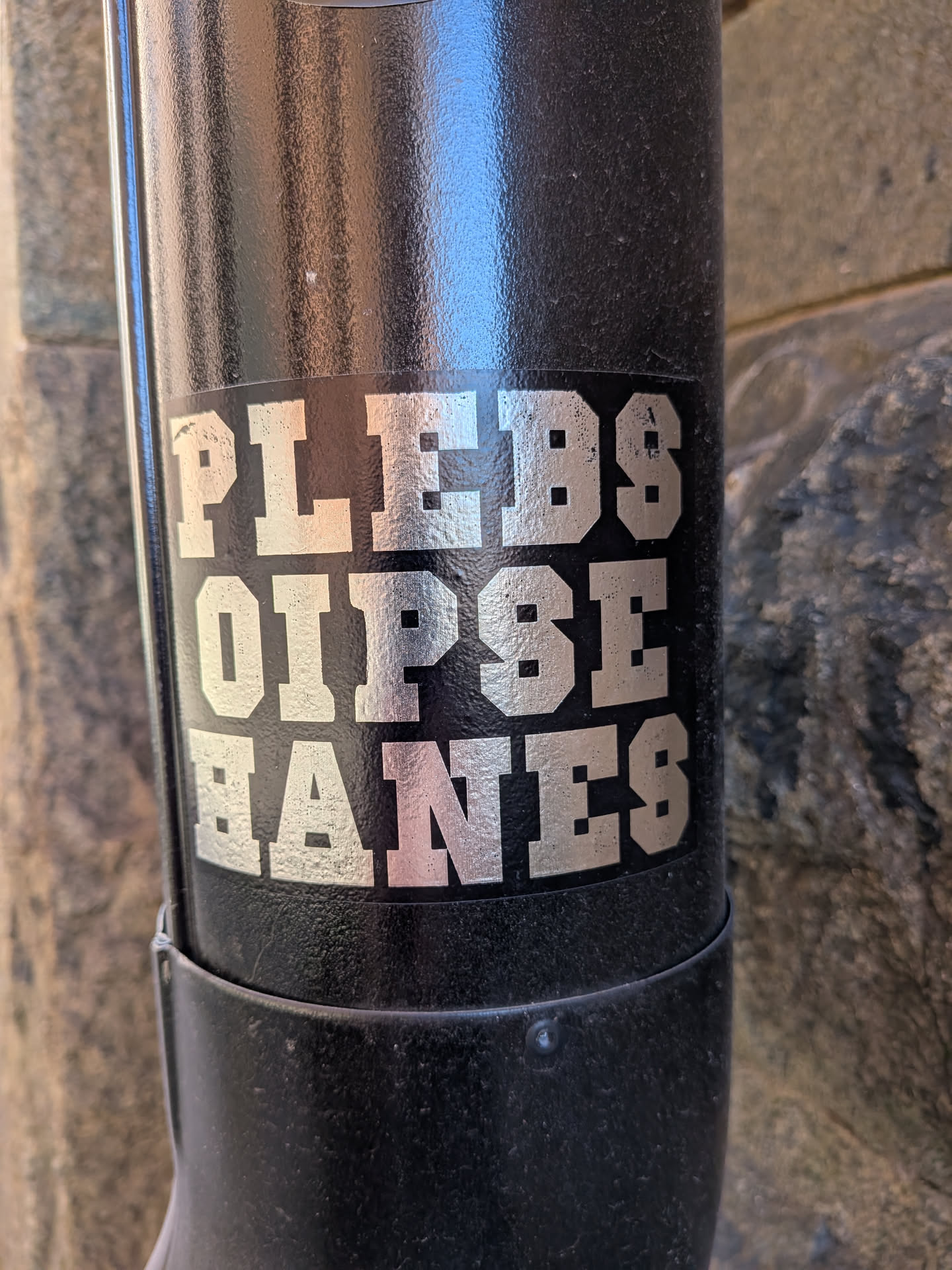 The sticker features the text "PLEBS OIPSE HANES" in large, bold, block-style letters. The letters are silver with a slightly distressed or textured finish, appearing almost metallic. They are arranged in three rows, with the first row containing "PLEBS", the second row "OIPSE", and the third row "HANES". The sticker has a black background, and its edges appear to be slightly worn or rounded, suggesting it is a peel-and-stick label applied to a curved black surface, possibly a pipe or a cylindrical object.