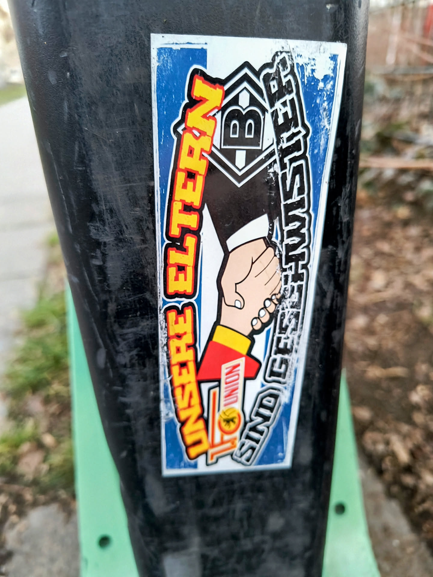 A sticker featuring a handshake, symbolizing the Union Berlin and Borussia Mönchengladbach football clubs' fans. On the left side, there's a hand in the Union Berlin team colors (red and yellow), and on the right, a hand with the logo of Borussia Mönchengladbach. The text reads "UNSERE ELTERN SIND GESCHWISTER" (our parents are siblings).