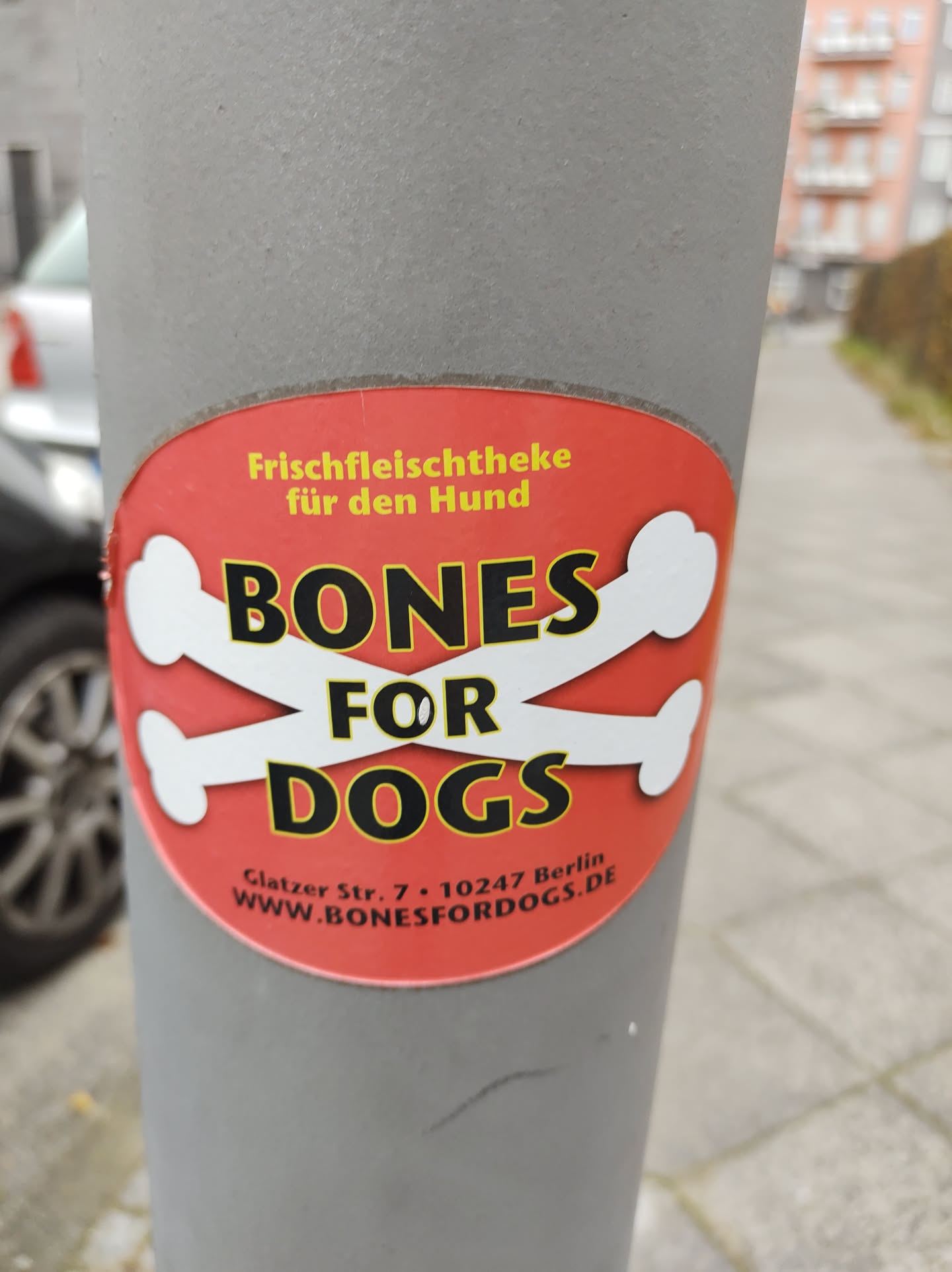 A red and white sticker advertising Bones for Dogs, a fresh meat shop for dogs located in Berlin, Germany.  The sticker features crossed bones and the shop's website address.