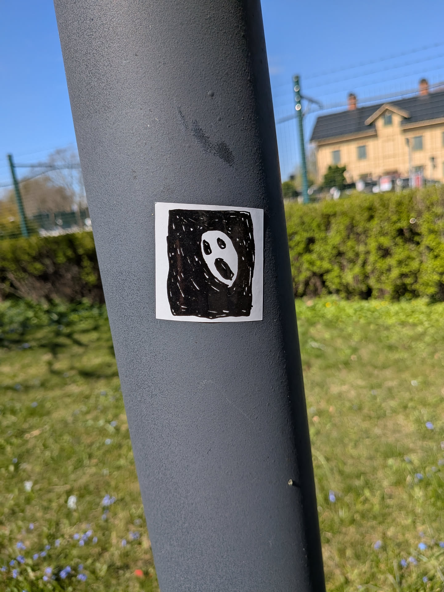 A square, white sticker featuring a hand-drawn, black and white illustration. The central image is a stylized, oval-shaped white face with two small, dark, oval eyes and a larger, open, oval mouth, giving the impression of a scream or shock. This white face is set against a roughly textured black background, created with visible white lines and strokes, suggesting a marker or ink drawing style. The overall aesthetic is raw and expressive, resembling a ghostly or distressed figure.