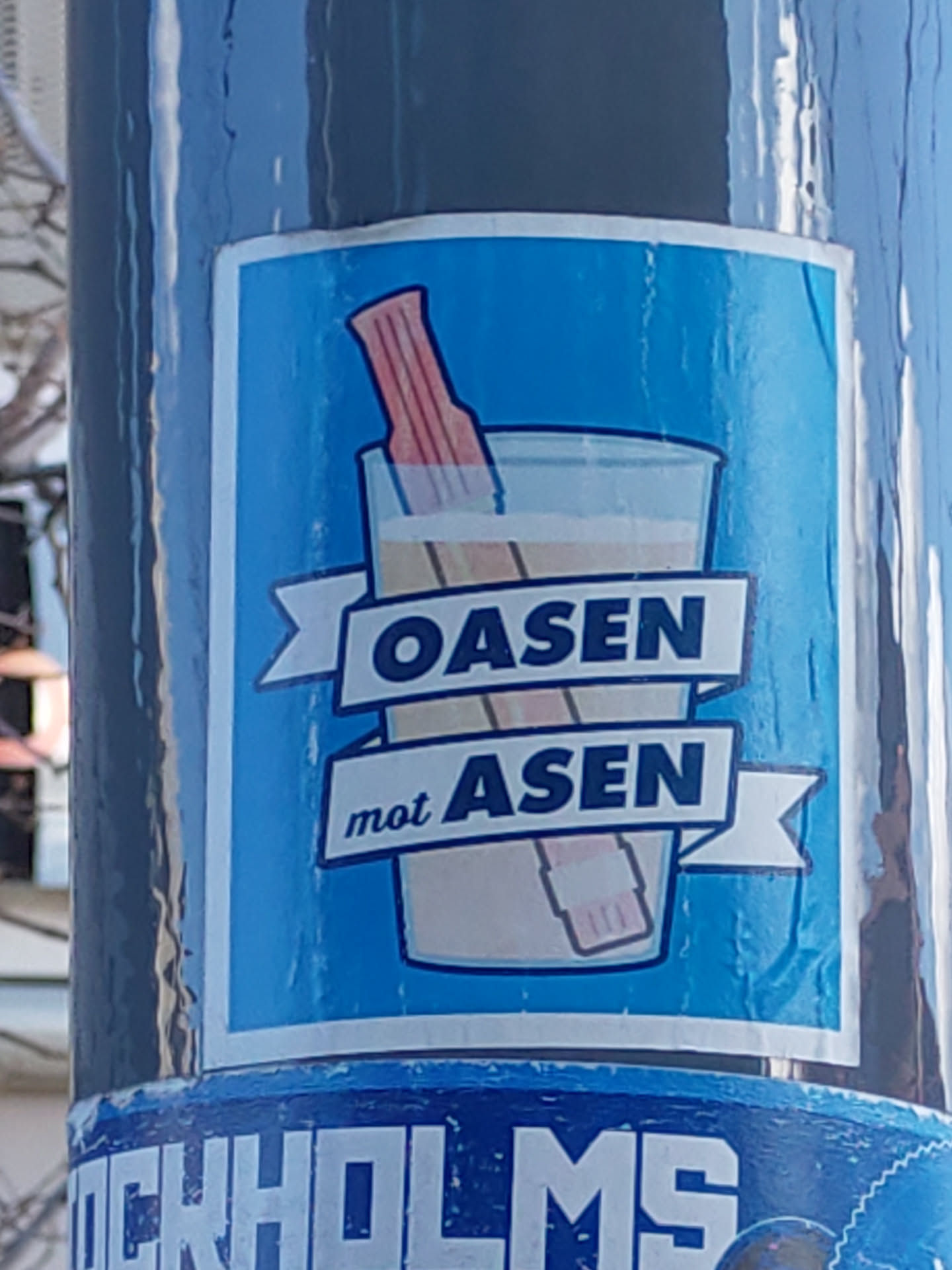A sticker depicting a pink object resembling a straw inside a glass of light-colored liquid.  The text "OASEN mot ASEN" is prominently displayed on a ribbon banner.