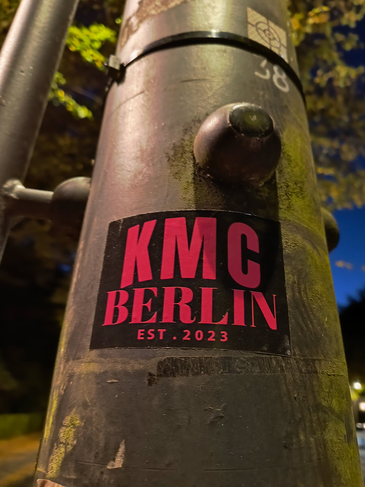 A black sticker with pink text on a lamppost. The text says KMC Berlin Est. 2023.