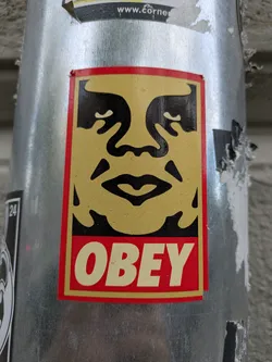A close-up shot of the classic OBEY sticker adhered to a metal pole. The sticker features the stylized face of Andre the Giant in black against a yellowish-tan background, bordered by a red frame. The word "OBEY" is printed in bold, capital letters at the bottom of the sticker. The pole shows signs of wear and other stickers, suggesting its urban environment.