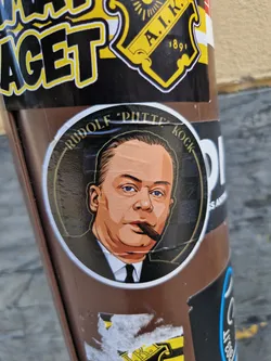 A round sticker featuring an illustration of Rudolf "Putte" Kock, a Swedish comedian and actor.  The illustration is in a vintage style, showing him smoking a cigar.