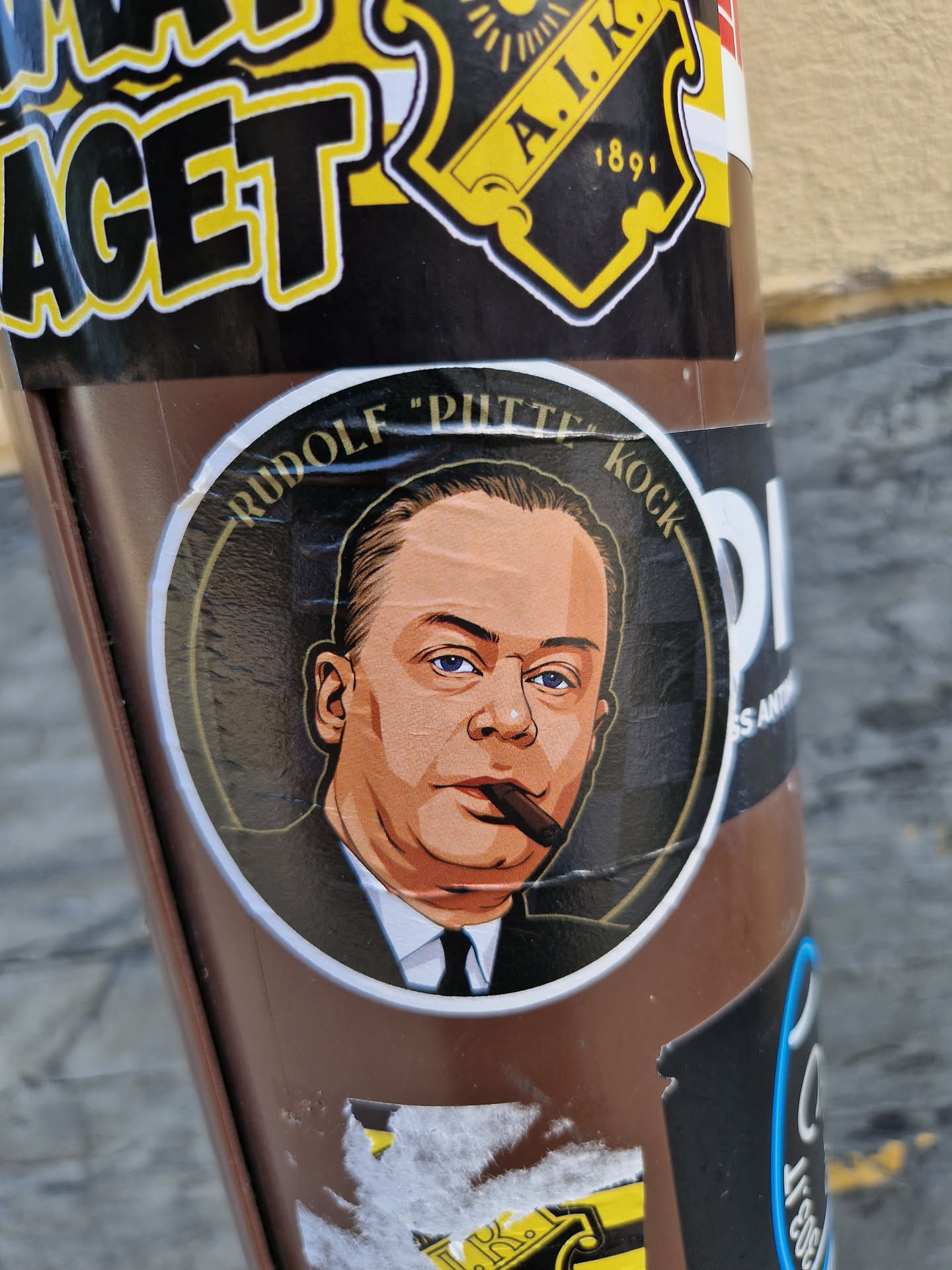 A round sticker featuring an illustration of Rudolf "Putte" Kock, a Swedish comedian and actor.  The illustration is in a vintage style, showing him smoking a cigar.