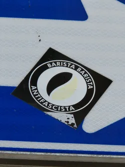 Black and white round sticker with the text "Barista Antifascista" written around a stylized coffee bean. The sticker is square and has some wear and tear.