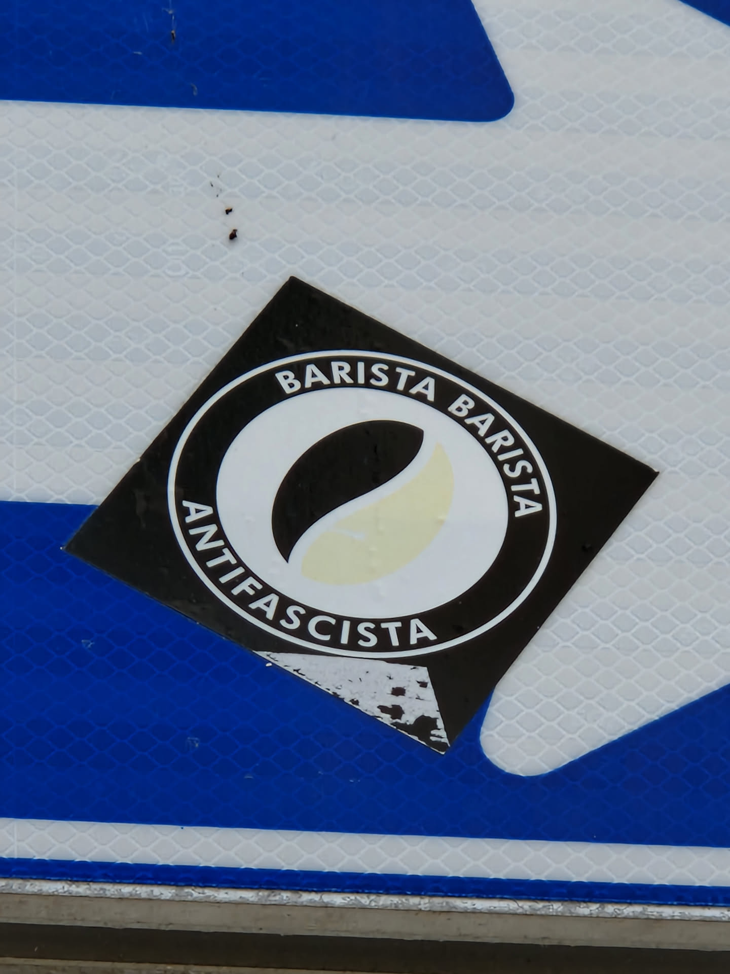 Black and white round sticker with the text "Barista Antifascista" written around a stylized coffee bean. The sticker is square and has some wear and tear.