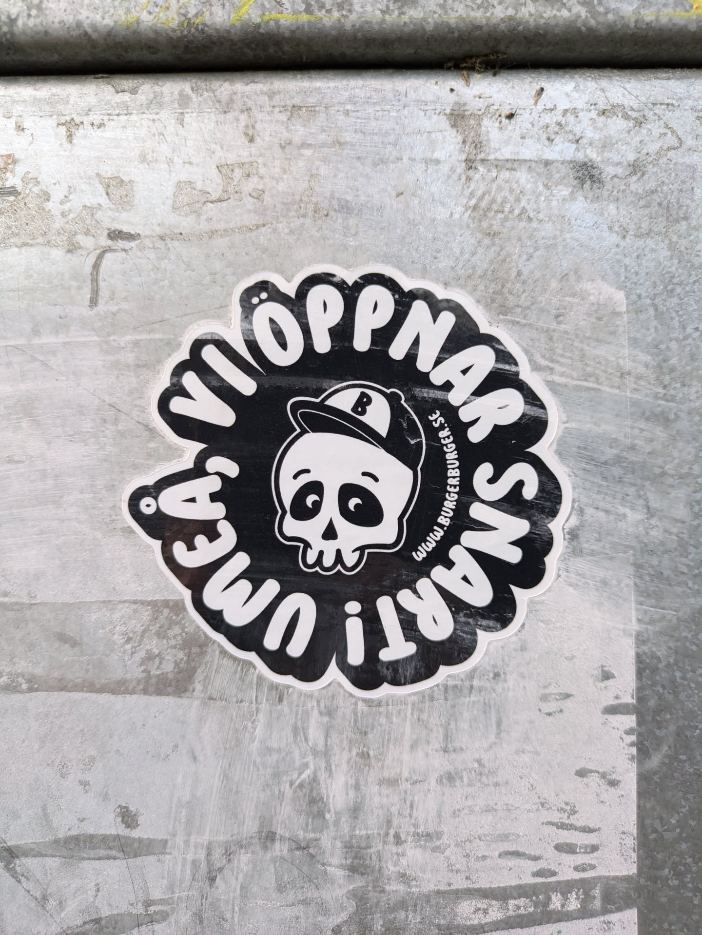 Black and white round sticker with a cartoon skull wearing a baseball cap in the center.  The text around the skull says  "VI ÖPPNAR SNART I UMEA", which translates from Swedish to "WE OPEN SOON IN UMEA". A small website address is visible but unreadable. 