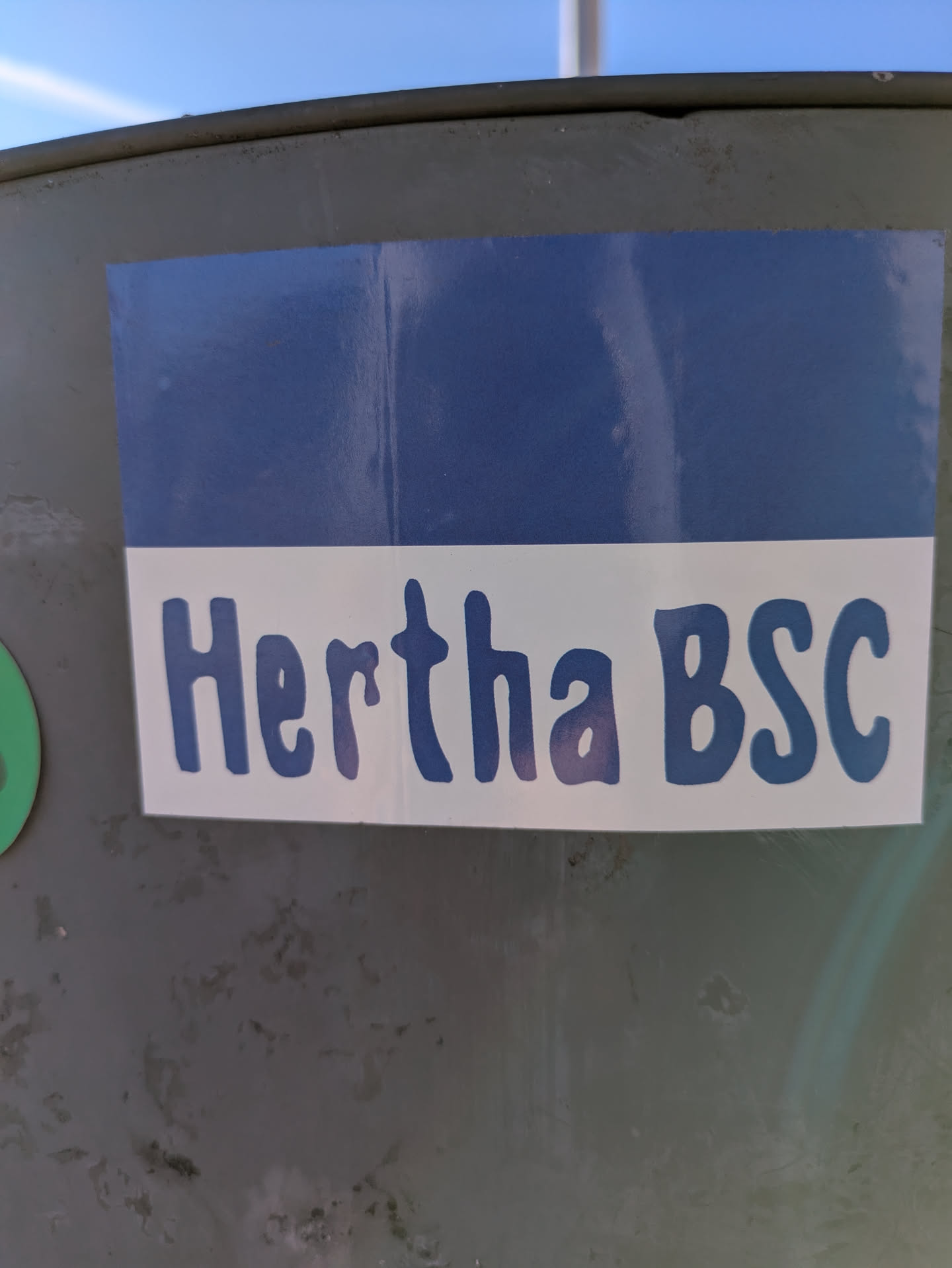 The sticker features the Hertha BSC logo with a blue and white color scheme. The text 'Hertha BSC' is prominently displayed in a rounded, blue font on a white background. The top portion of the sticker is a solid blue. The sticker is affixed to a slightly weathered, grey metal surface.