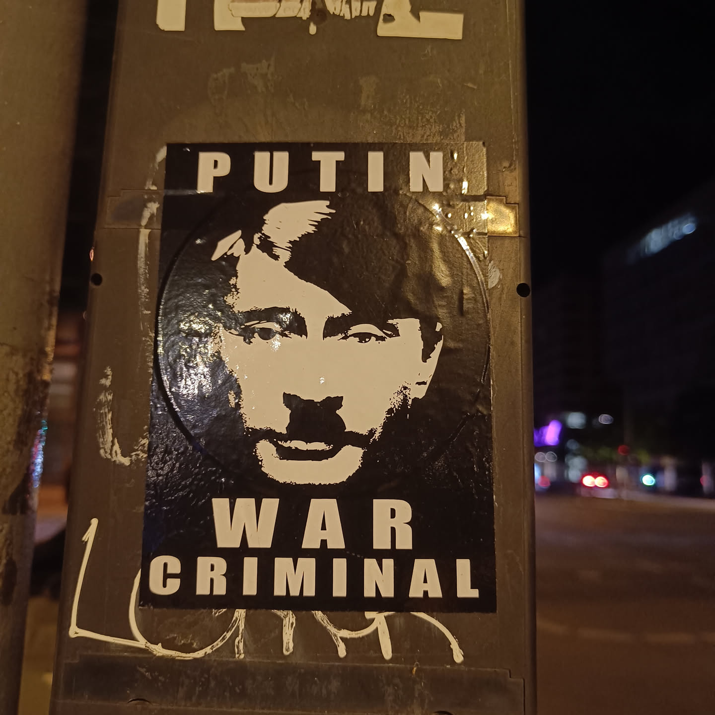 A black and white sticker depicting Vladimir Putin's face with the text 'PUTIN WAR CRIMINAL'. The sticker is affixed to a dark-colored pole at night.