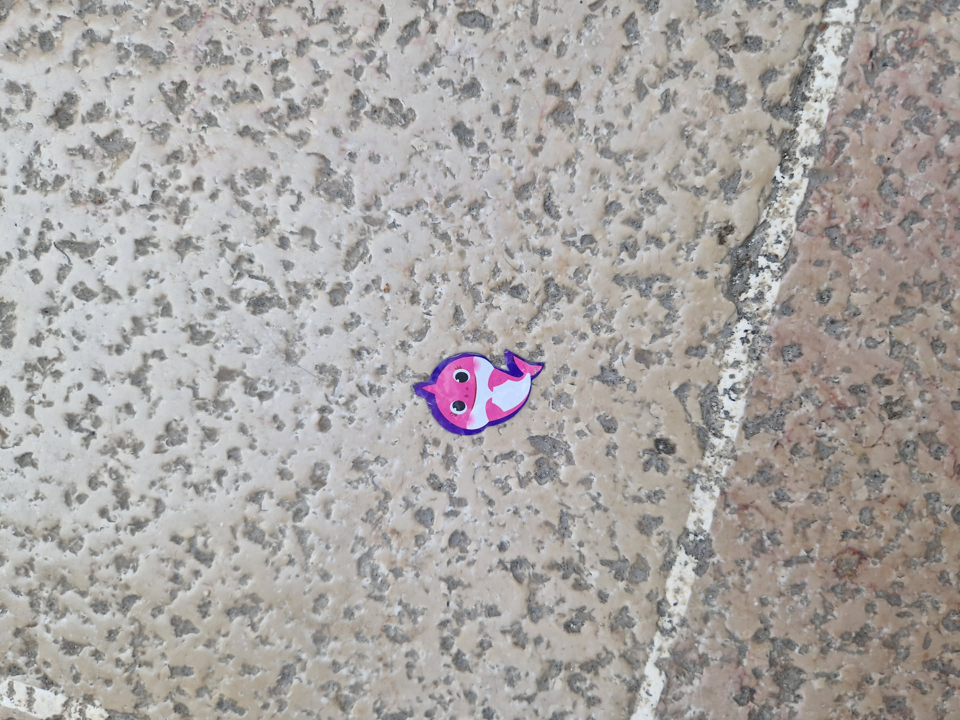 A small pink baby shark sticker is placed on a textured beige ground. The sticker features a cartoonish baby shark with a simple design.