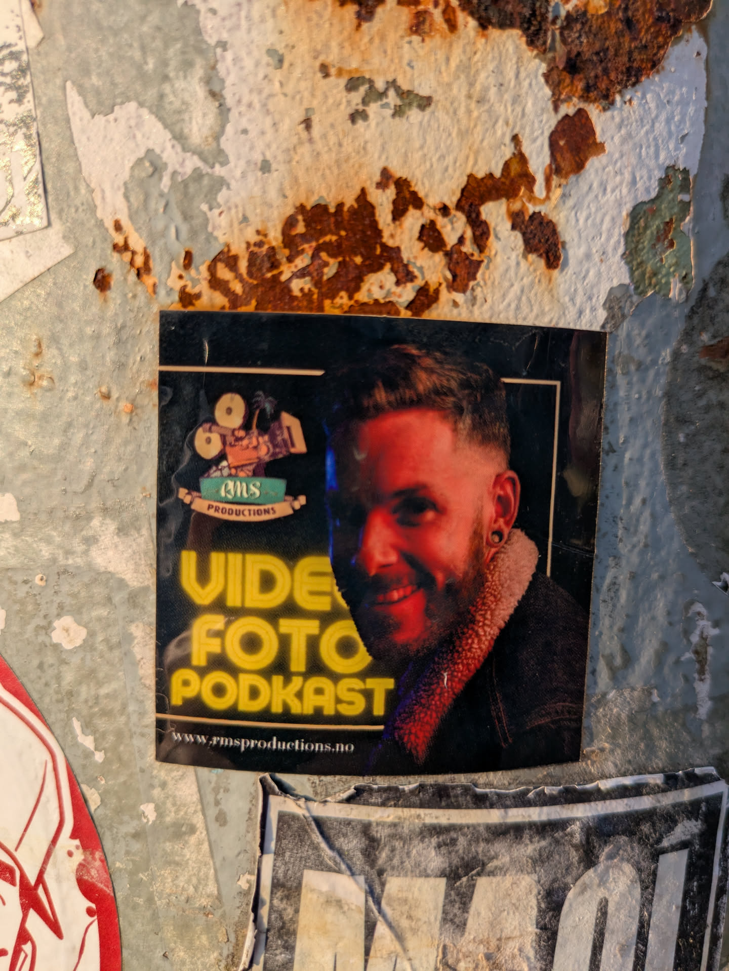 A square-shaped sticker with a black background, featuring a smiling man with a beard, short hair, and ear tunnels. His face is illuminated with a reddish hue, possibly from ambient lighting. He is wearing a dark jacket with a textured, possibly fur or shearling, collar. In the top left corner, there's a logo with film cameras and the text "RMS PRODUCTIONS". Below the man's portrait, large yellow text reads "VIDE FOTO PODKAST". At the very bottom, a website URL "www.rmsproductions.no" is visible.