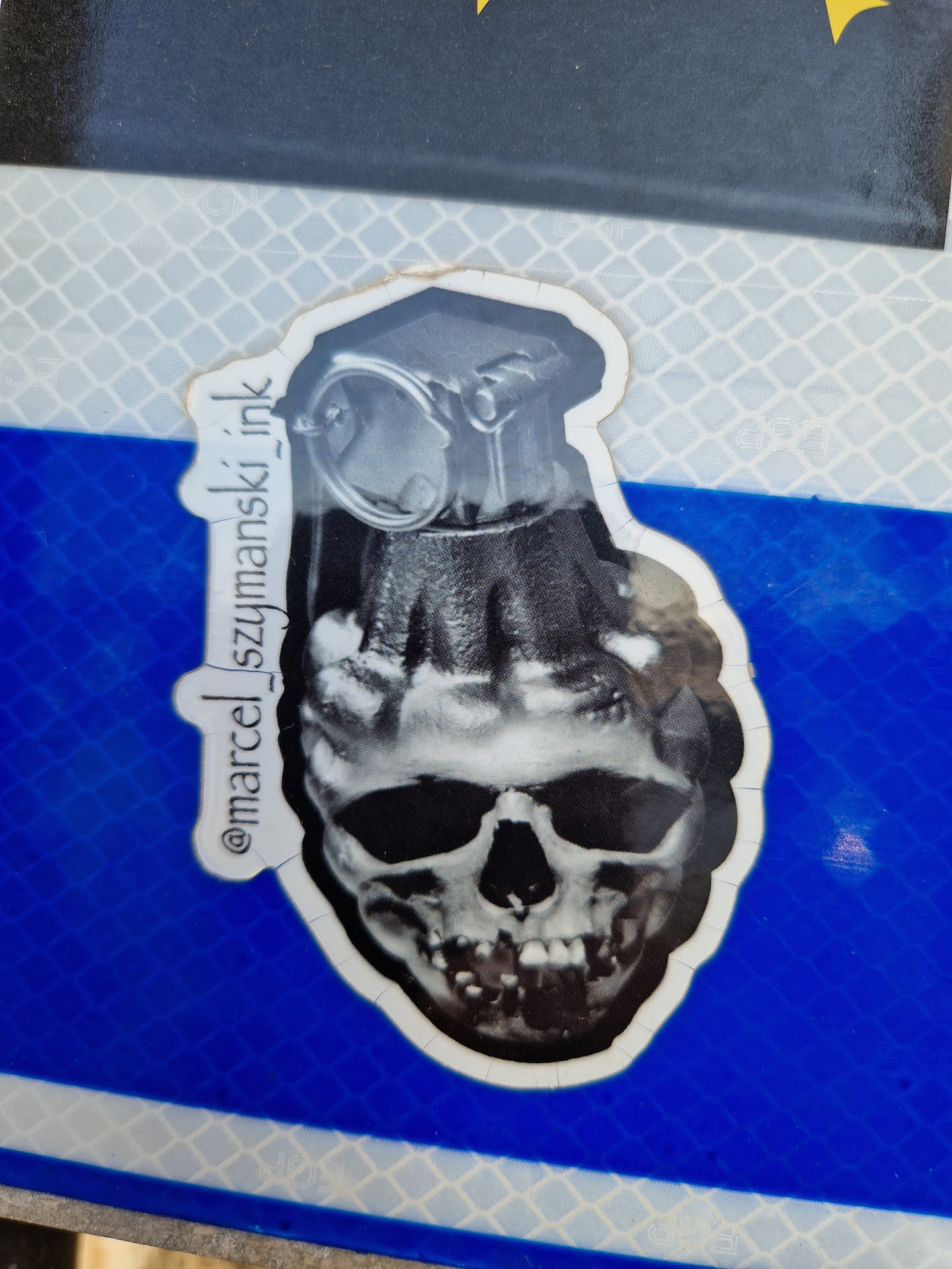 Black and white sticker of a skull with a grenade on top. The artist's Instagram handle is visible on the sticker.