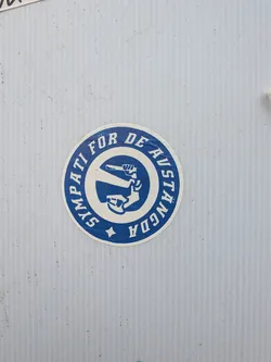 A circular sticker featuring a two-tone design in deep blue and off-white. The outer ring is blue with the text 'SYMPATI FÖR DE AVSTÄNGDA' in white capital letters, encircling the central image. A small white star is positioned at the bottom of this outer ring. The inner circle has an off-white background and displays a stylized blue graphic of a clenched fist holding a wrench, angled diagonally. The sticker shows signs of wear and age, with some dirt and minor scuffs, indicating it has been affixed for some time.