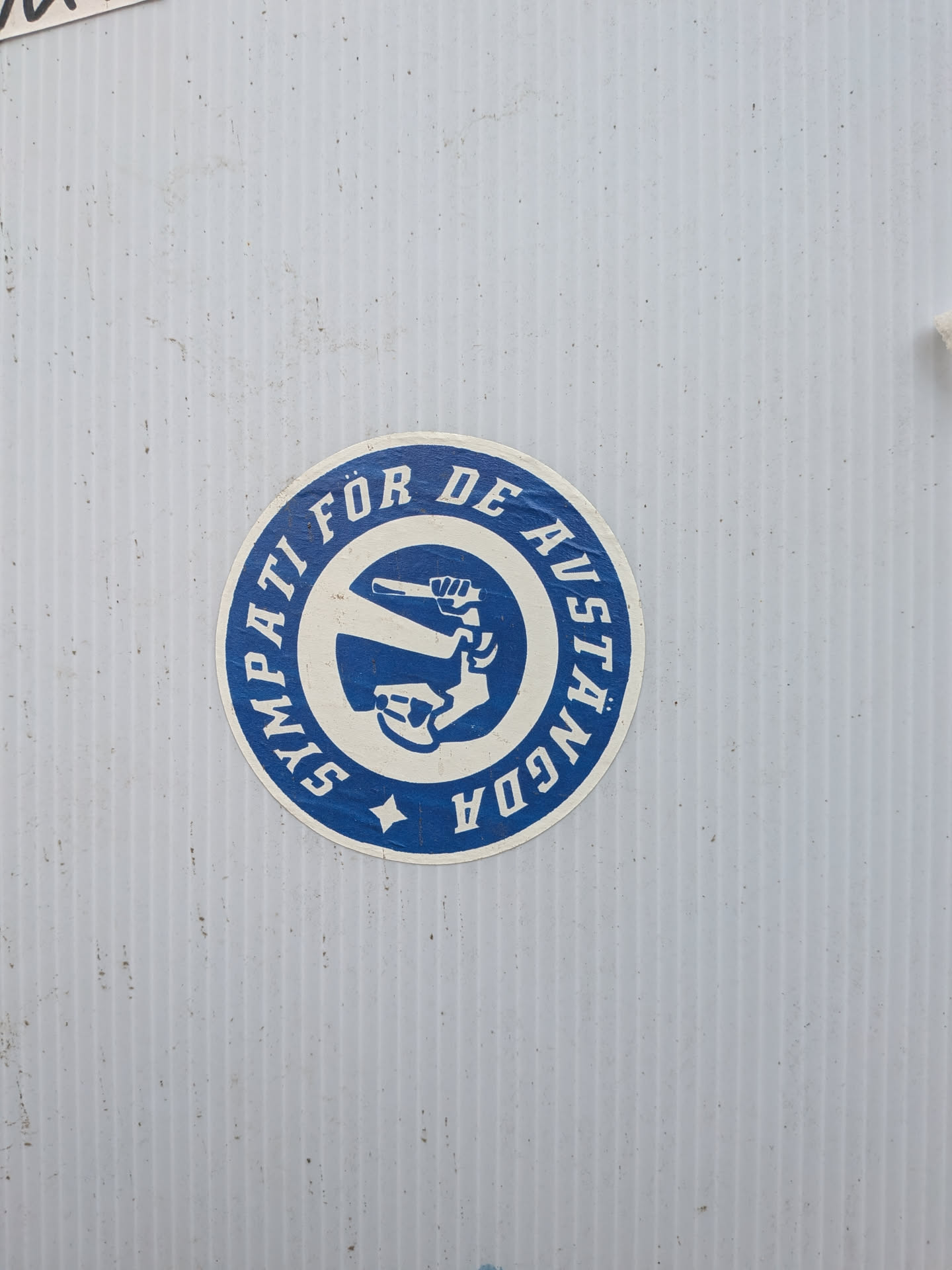A circular sticker featuring a two-tone design in deep blue and off-white. The outer ring is blue with the text 'SYMPATI FÖR DE AVSTÄNGDA' in white capital letters, encircling the central image. A small white star is positioned at the bottom of this outer ring. The inner circle has an off-white background and displays a stylized blue graphic of a clenched fist holding a wrench, angled diagonally. The sticker shows signs of wear and age, with some dirt and minor scuffs, indicating it has been affixed for some time.