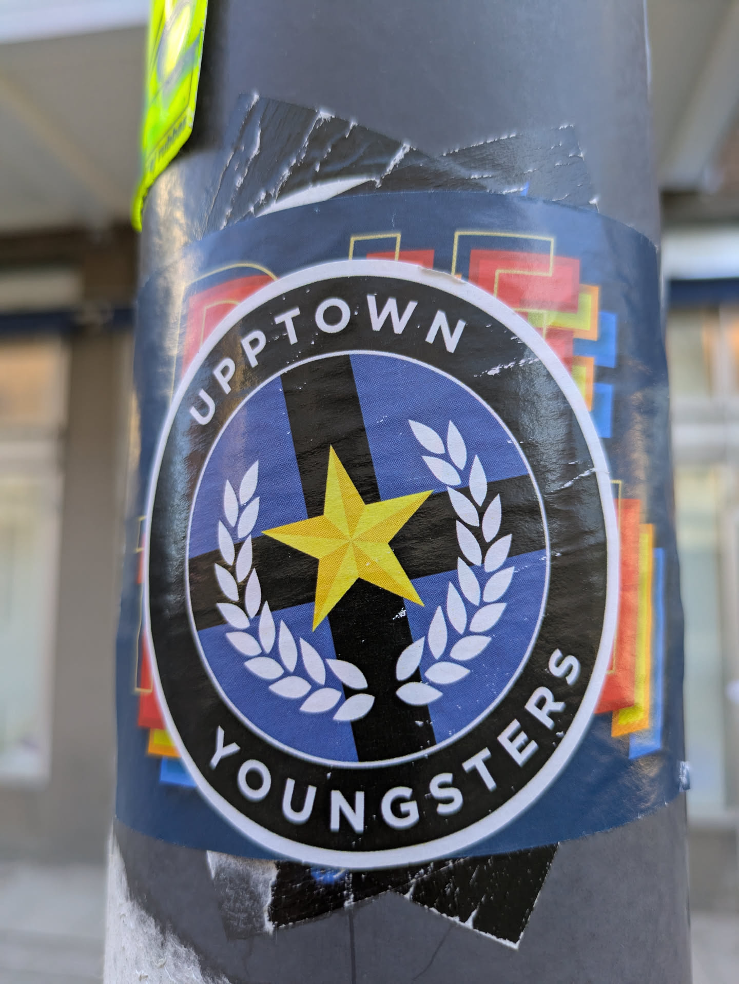 The sticker features the text "UPPTOWN YOUNGSTERS" in a circular arrangement around an inner circle. The inner circle has a blue and black cross design, overlaid with a yellow star and surrounded by laurel branches. The outer edges of the sticker has colorful geometric shapes.