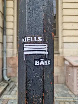A black and white sticker featuring the text "KJELLS BÄNK" and an illustration of a bench. The sticker is applied to a weathered dark gray metal pole. 