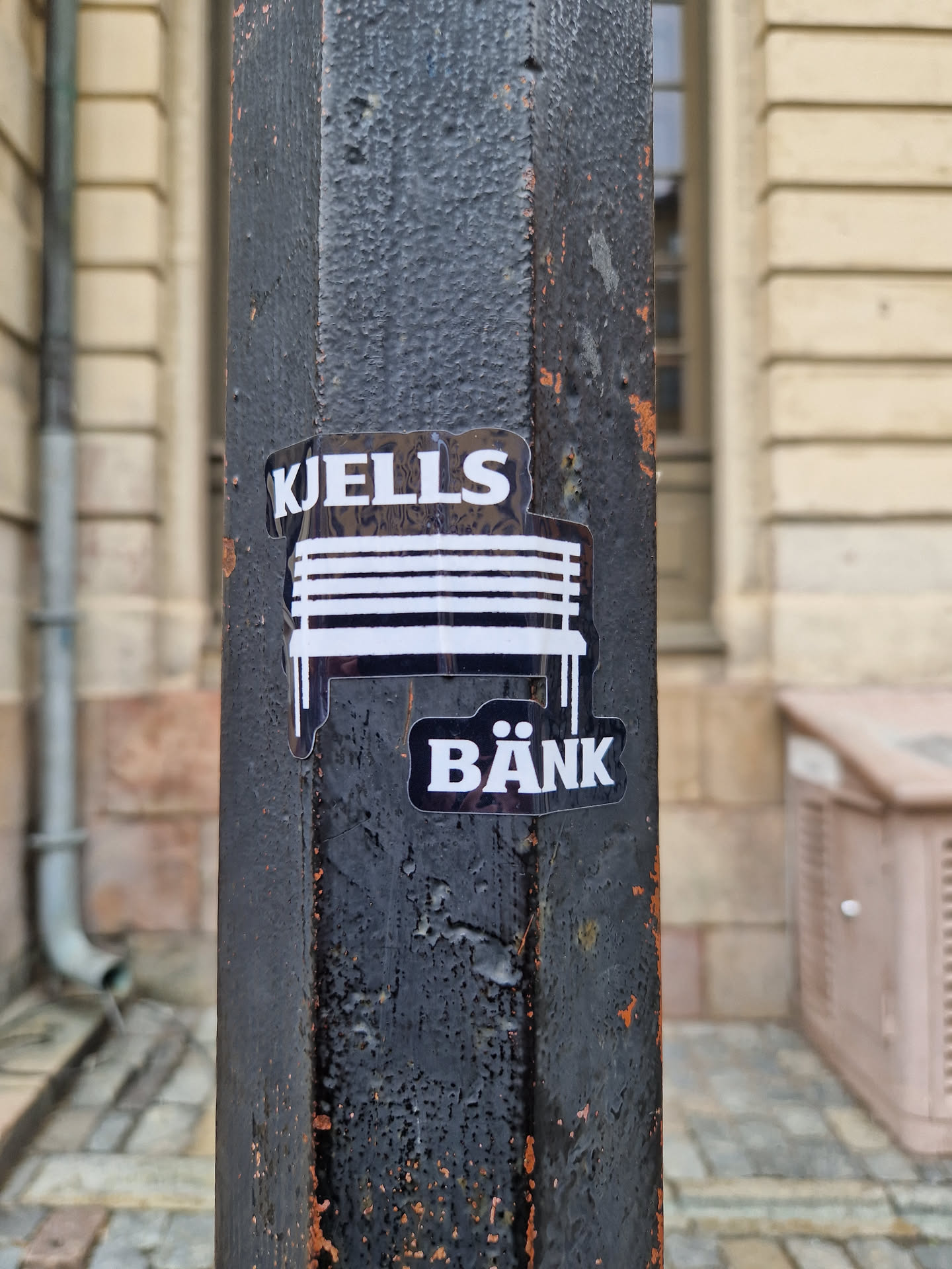A black and white sticker featuring the text "KJELLS BÄNK" and an illustration of a bench. The sticker is applied to a weathered dark gray metal pole. 
