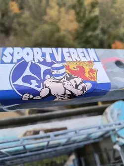 A sticker featuring a muscular figure wearing a mask, along with a logo that reads "SPORTVEREIN". The design is colorful and visually striking. The sticker is affixed to a metal surface outdoors.