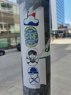 The sticker features a vertical arrangement of four distinct images. Topmost is a stylized flag, followed by a circular emblem that reads "1905 G.I.F. STUDS". Below is a skull and crossbones motif, and at the bottom is a graphic resembling a hat. The stickers are layered on a metal pole, showing signs of wear and tear. 