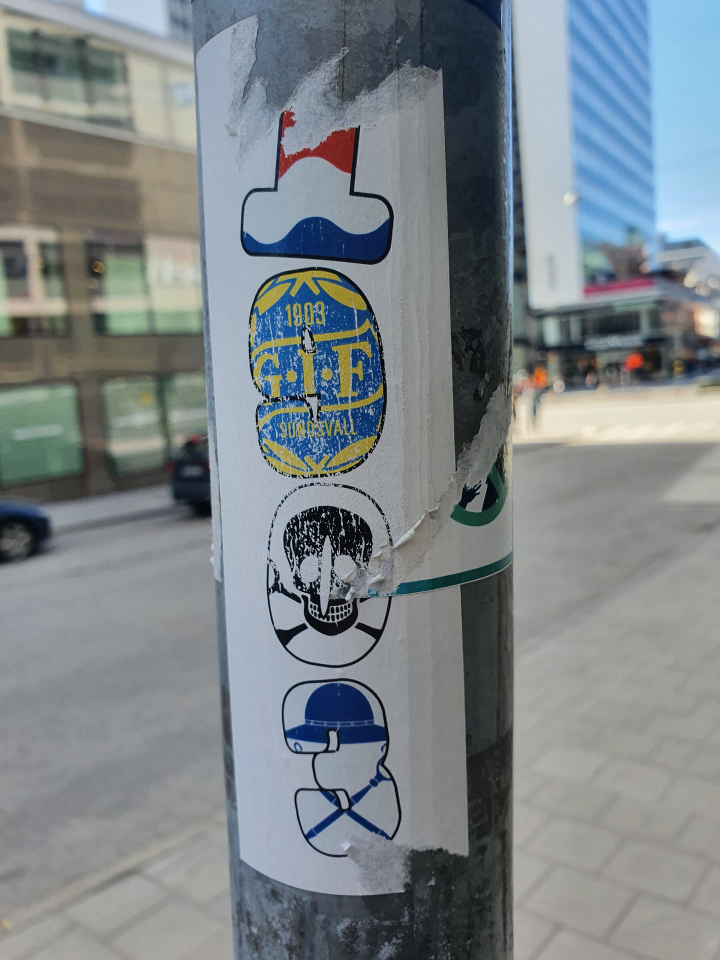 The sticker features a vertical arrangement of four distinct images. Topmost is a stylized flag, followed by a circular emblem that reads "1905 G.I.F. STUDS". Below is a skull and crossbones motif, and at the bottom is a graphic resembling a hat. The stickers are layered on a metal pole, showing signs of wear and tear. 