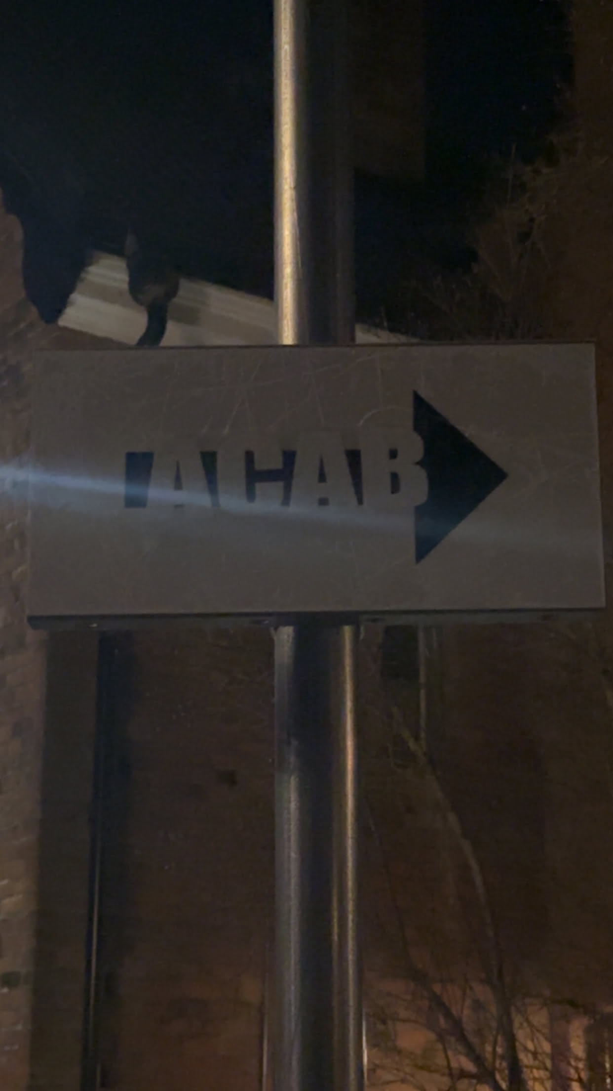 A rectangular sign or sticker, horizontally oriented, featuring the acronym 'ACAB' in bold, black, sans-serif capital letters. Immediately following the text is a solid black arrow pointing to the right. The background of the sign is a light grey or off-white. The surface shows minor scratches and wear, suggesting it is a physical object attached to a metal pole. The overall design is simple, direct, and monochrome.