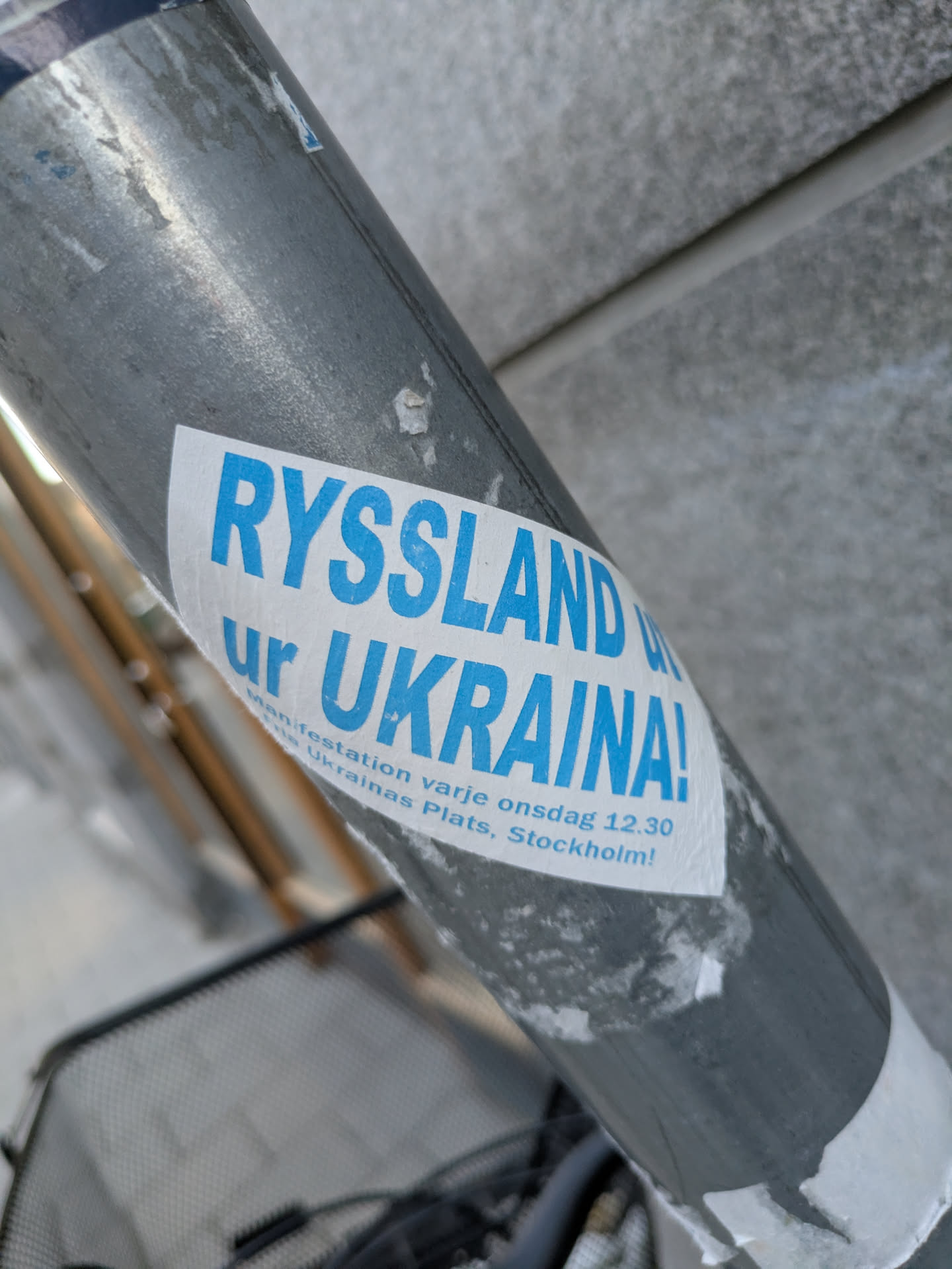 A white sticker with blue text reading "RYSSLAND UT UR UKRAINA!" is adhered to a gray metal pole, possibly a lamppost or signpost. Below the main text, smaller text states "Manifestation varje onsdag 12.30 Ukrainas Plats, Stockholm!" The sticker seems to be advocating for Russia to leave Ukraine and promoting a weekly manifestation in Stockholm.