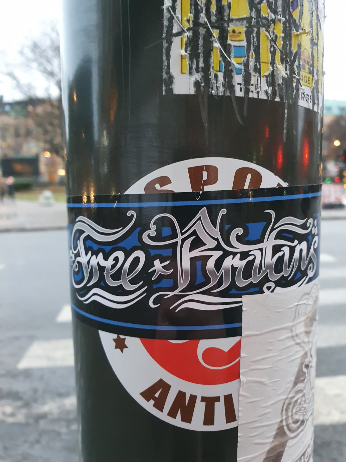 A sticker with the text "Free Furryans" in silver stylized lettering on a dark background with a blue stripe at the bottom.  The sticker is affixed to a pole, among other stickers.