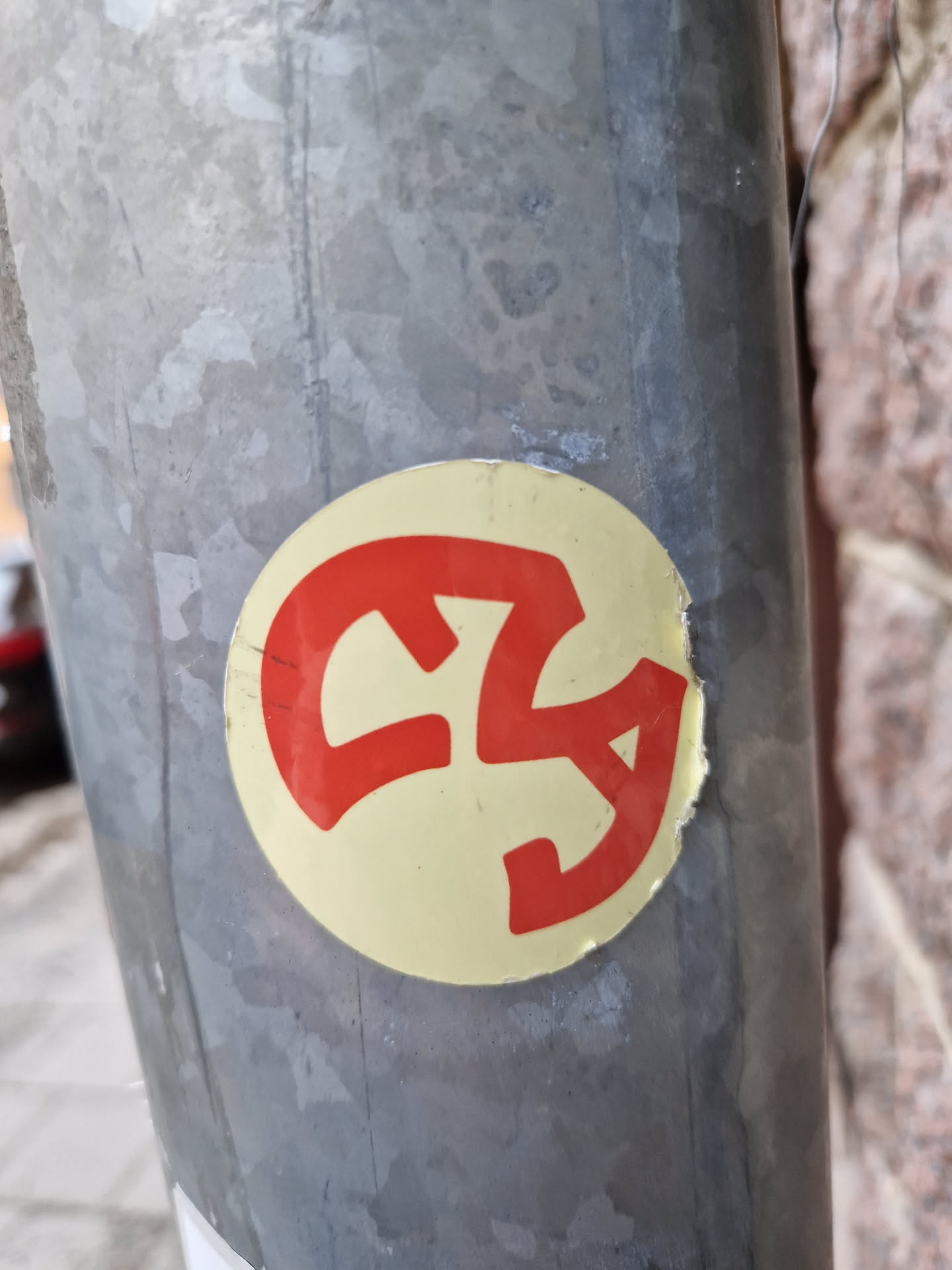 A round sticker with a red abstract design on a pale yellow background. The design is somewhat reminiscent of a stylized logo or symbol.  The sticker is affixed to a grey metal pole. A portion of the sticker appears to be peeled or damaged.