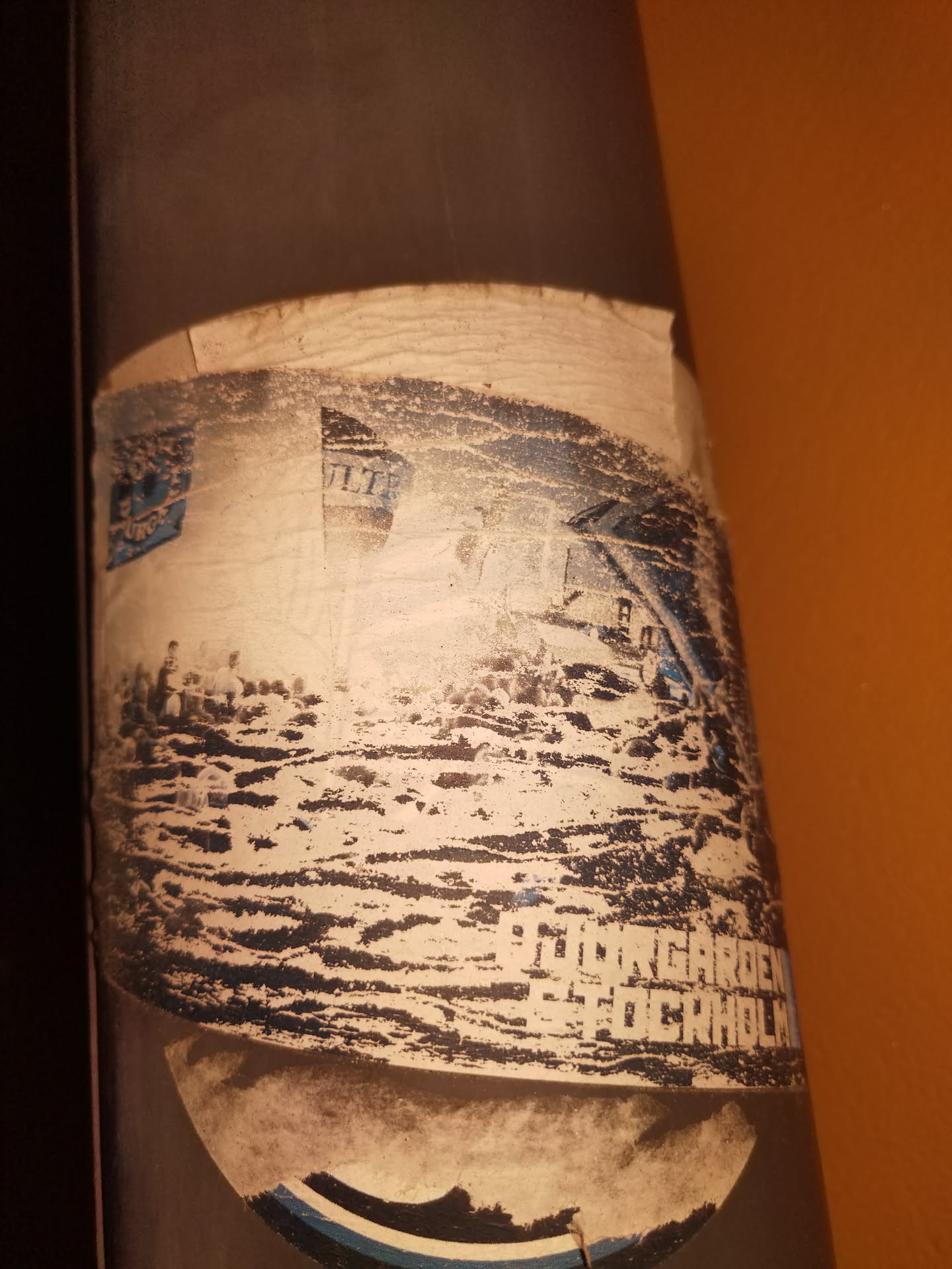 This sticker is affixed to a pole and shows significant wear and tear. It depicts a view of Djurgården, an island in Stockholm, Sweden. The text 'DJURGÅRDEN STOCKHOLM' is partially visible at the bottom. The image is somewhat faded and partially obscured by damage to the sticker.