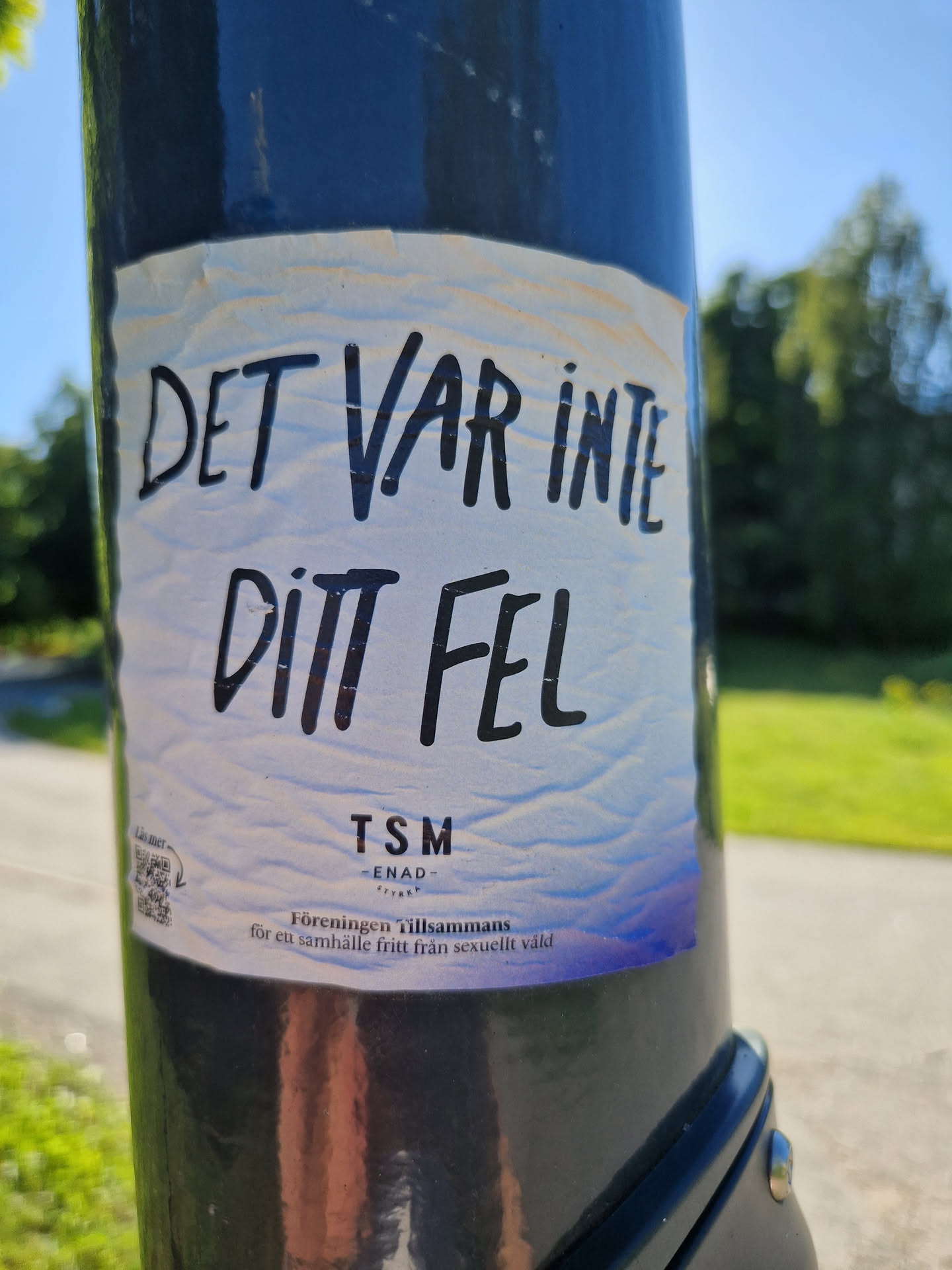 A sticker placed on a lamppost advocating against sexual violence.  The sticker features text in Swedish that translates to "It wasn't your fault."  It also includes the name of an organization, TSM - ENAD (likely an abbreviation). A QR code is present but the URL it directs to could not be determined. 