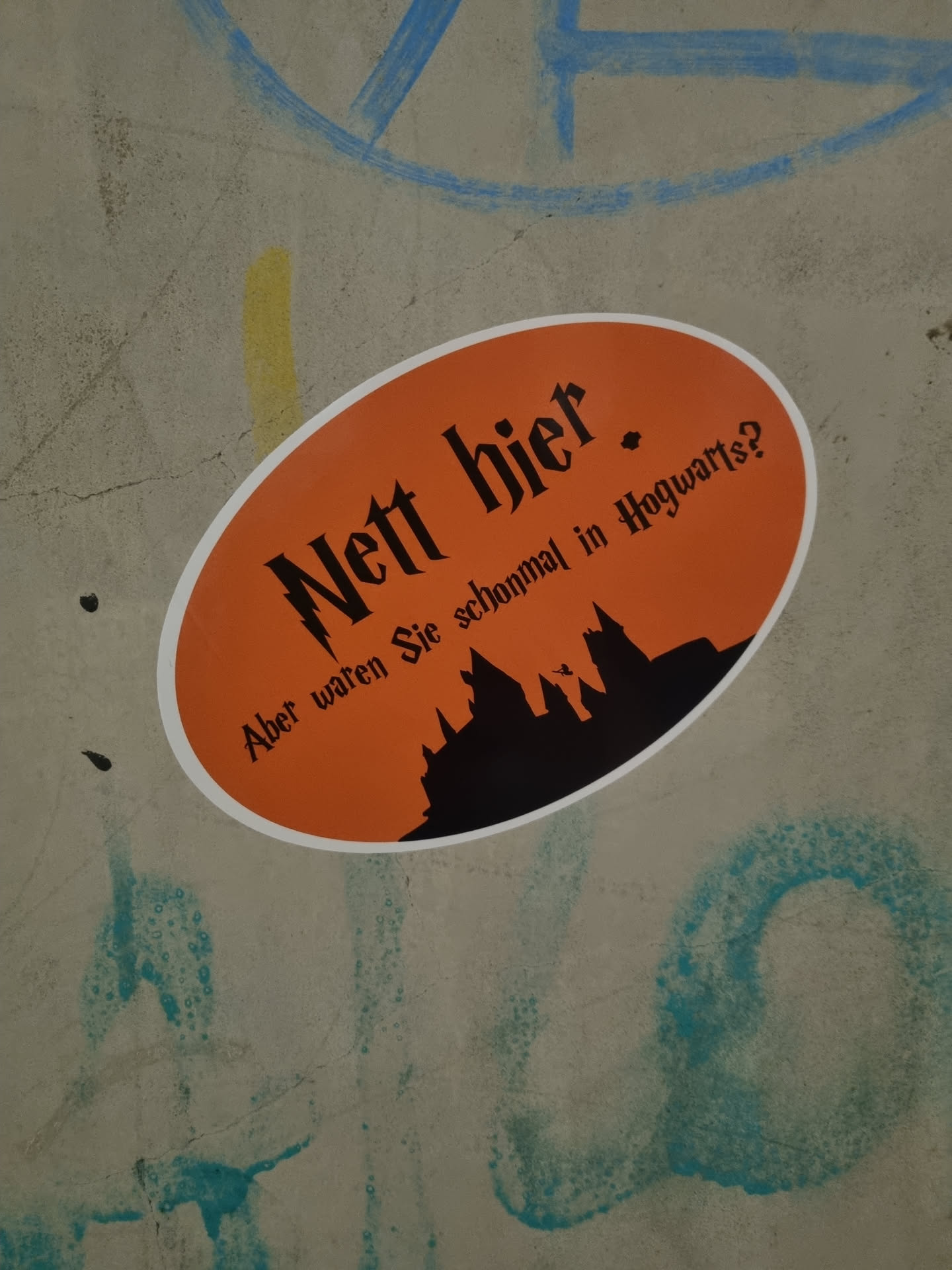 An orange oval sticker featuring a silhouette of Hogwarts castle with the text "Nett hier. Aber waren Sie schonmal in Hogwarts?" which translates to "Nice here. But have you ever been to Hogwarts?" in German. The sticker is affixed to a grey concrete surface.
