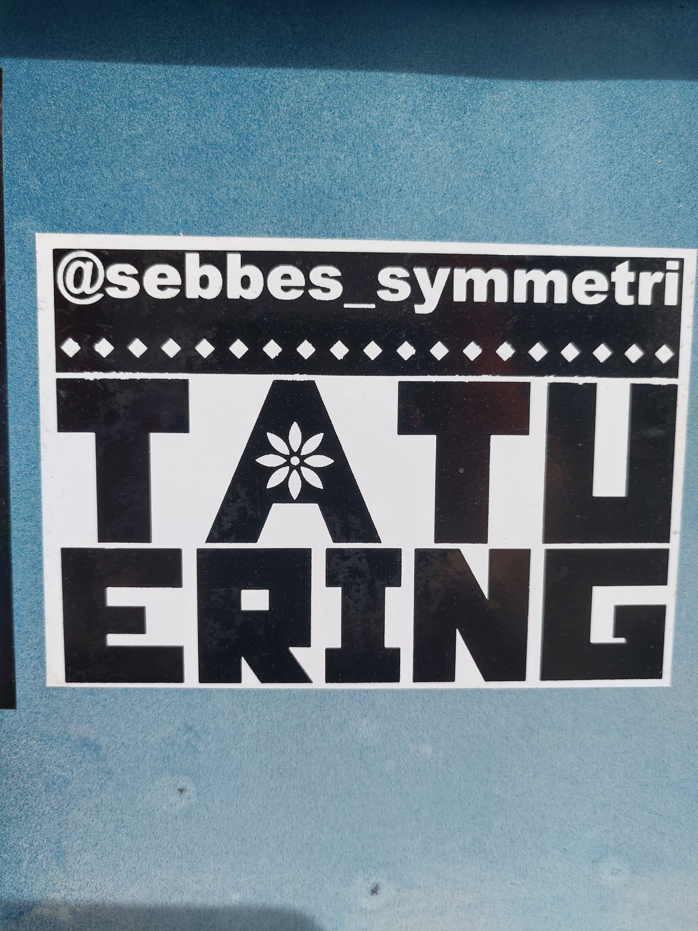 Black and white sticker with the word TATUERING and a small flower in the center of the A. The Instagram handle @sebbes_symmetri is at the top of the sticker.