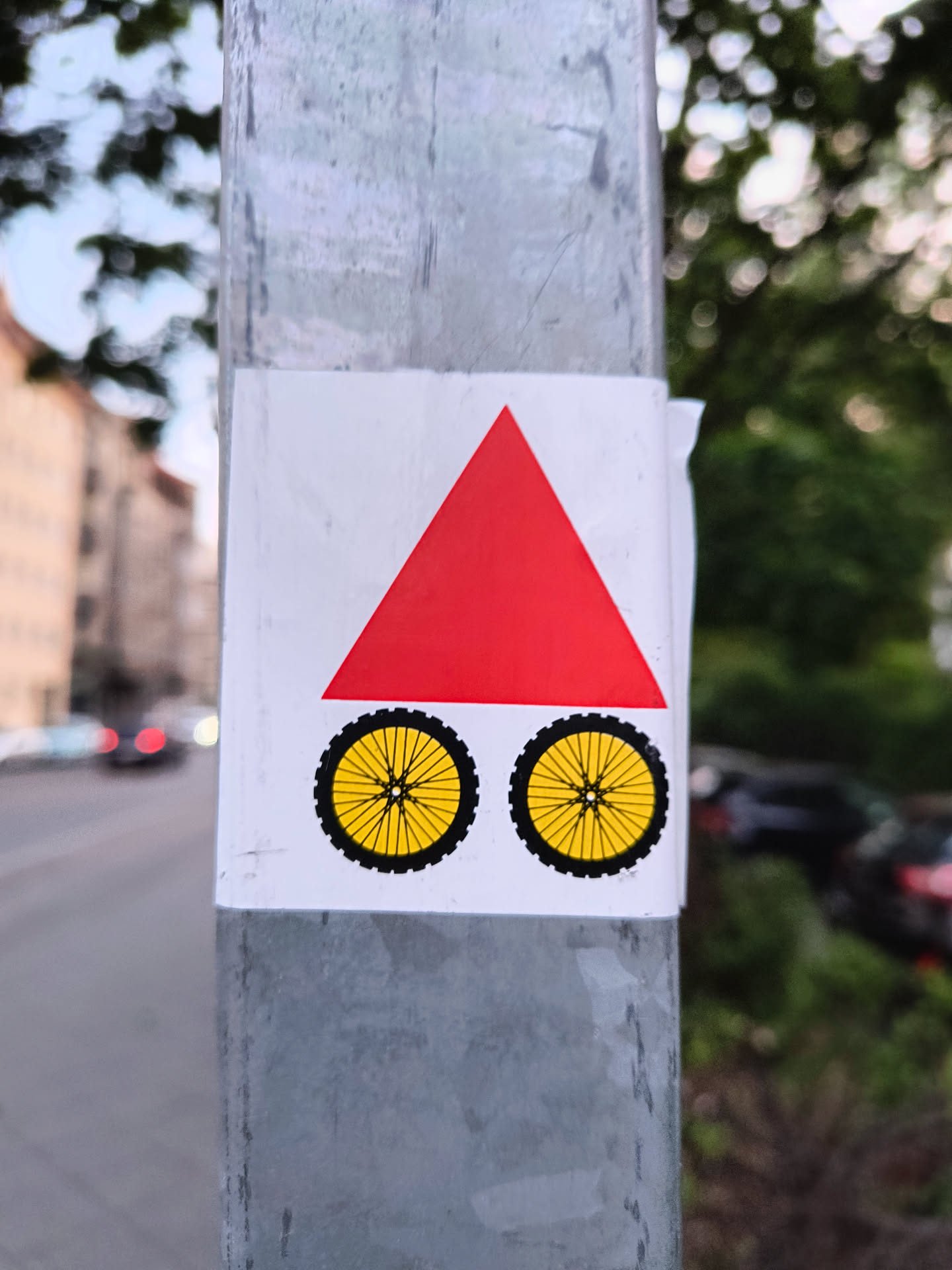This sticker features a red triangle sitting atop two bicycle wheels. The triangle is equilateral and solid red. The wheels are yellow with black tires and detailed spokes. It's pasted on a grey metal pole.