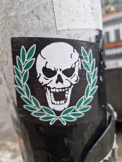 A sticker featuring a skull design with a laurel wreath around it. The skull is white, and the wreath is green on a black background. The sticker appears to be placed on a pole or similar surface outdoors.