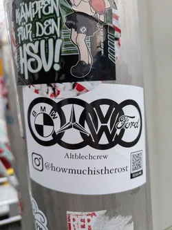 A white, rectangular sticker affixed to a pole, showcasing a prominent black graphic of five well-known car manufacturer logos – BMW, Mercedes-Benz (star), Audi (four rings), Volkswagen (VW), and Ford – creatively interwoven into a single design. Below this central graphic, the text 'Altblechcrew' is printed. Further down, an Instagram icon precedes the handle '@howmuchistherost'. In the bottom right corner, a QR code is visible with the words 'SCAN ME' directly beneath it.