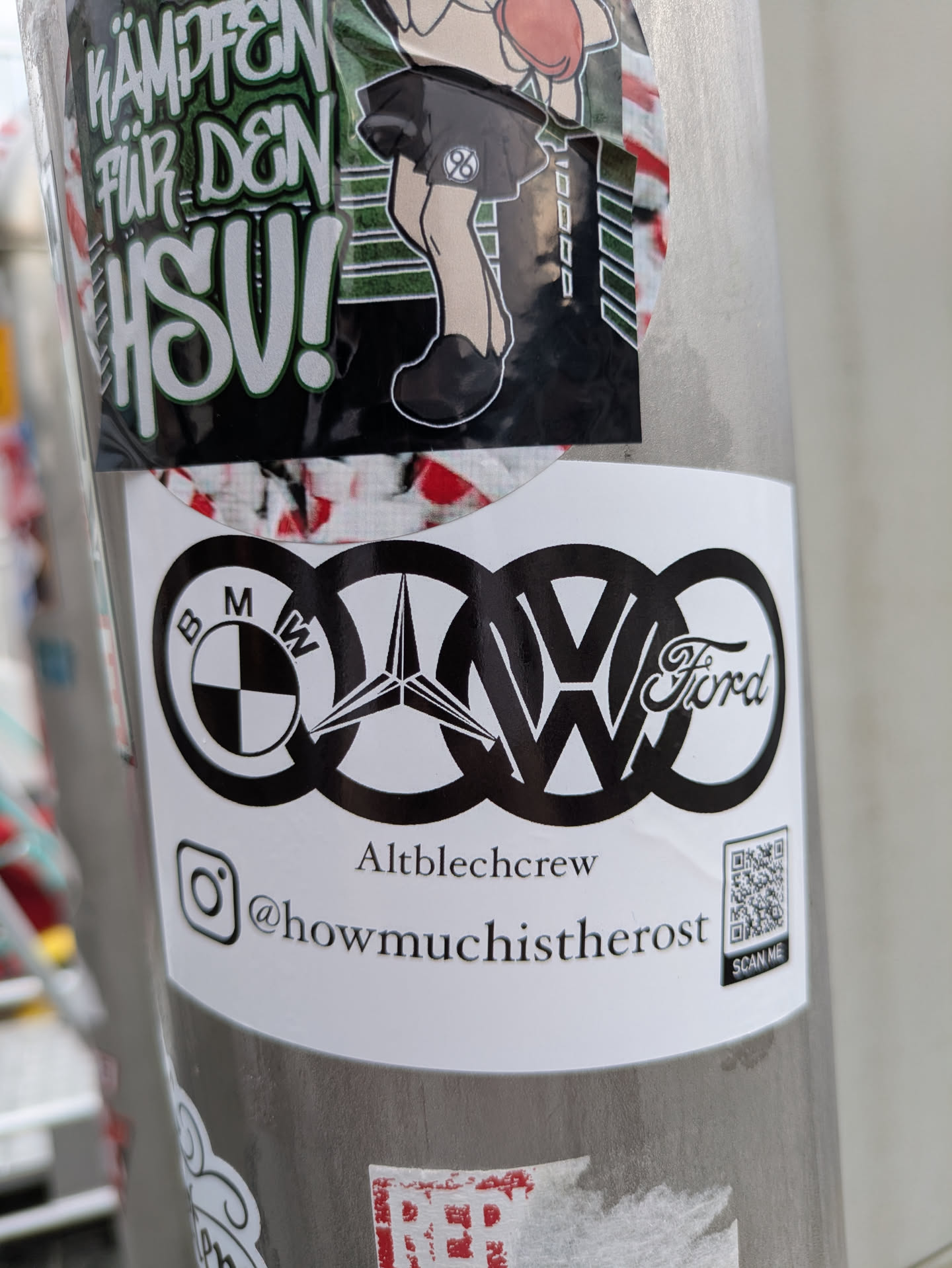 A white, rectangular sticker affixed to a pole, showcasing a prominent black graphic of five well-known car manufacturer logos – BMW, Mercedes-Benz (star), Audi (four rings), Volkswagen (VW), and Ford – creatively interwoven into a single design. Below this central graphic, the text 'Altblechcrew' is printed. Further down, an Instagram icon precedes the handle '@howmuchistherost'. In the bottom right corner, a QR code is visible with the words 'SCAN ME' directly beneath it.