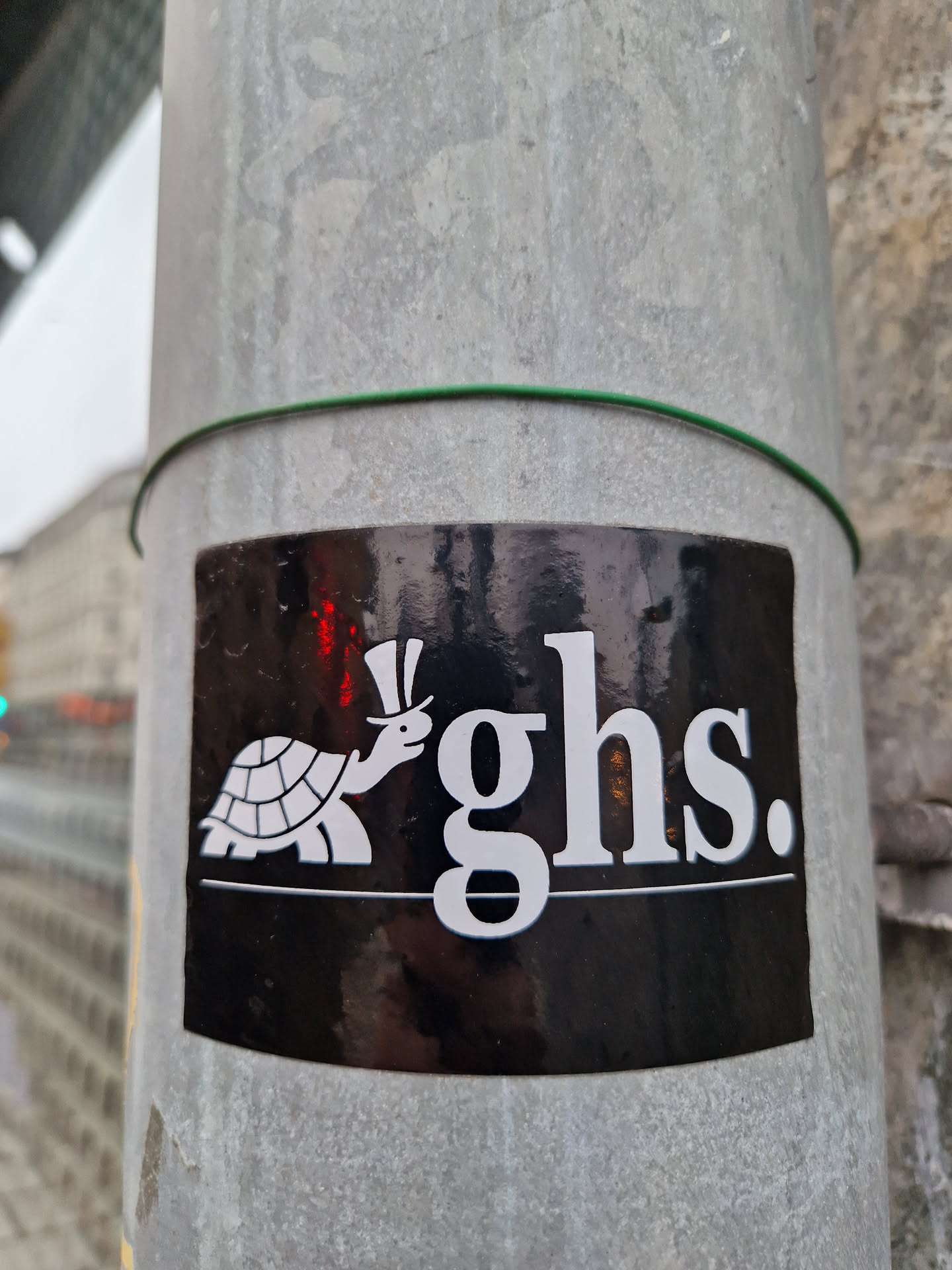 A black square sticker with a white illustration of a turtle wearing a top hat, accompanied by the stylized letters 'ghs.' The sticker is affixed to a grey metal pole.