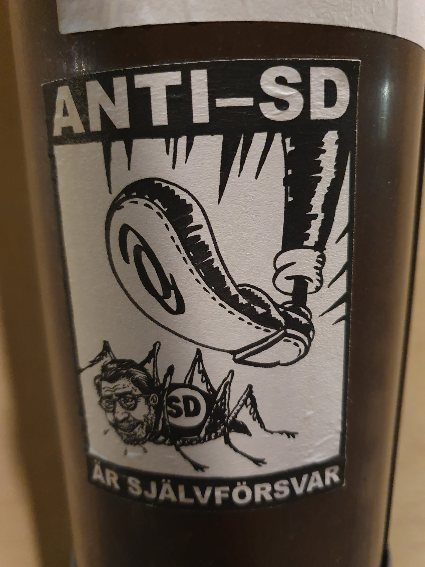 Black and white sticker with an illustration of a foot crushing a cricket with a man's face. The cricket has the letters "SD" on its back. The text on the sticker says "ANTI-SD ÄR SJÄLVFÖRSVAR".