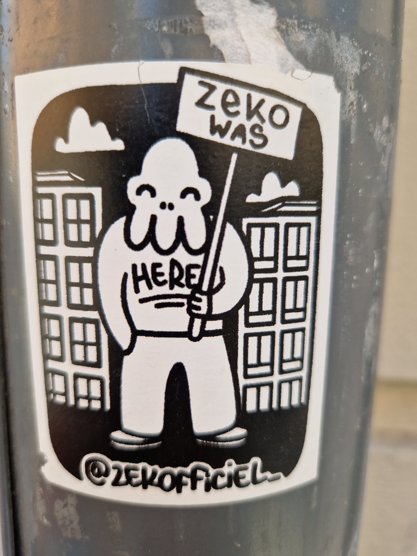 Black and white sticker featuring a cartoon character holding a sign that says "ZeKo was here". The character is standing in front of buildings. The sticker includes the artist's Instagram handle.
