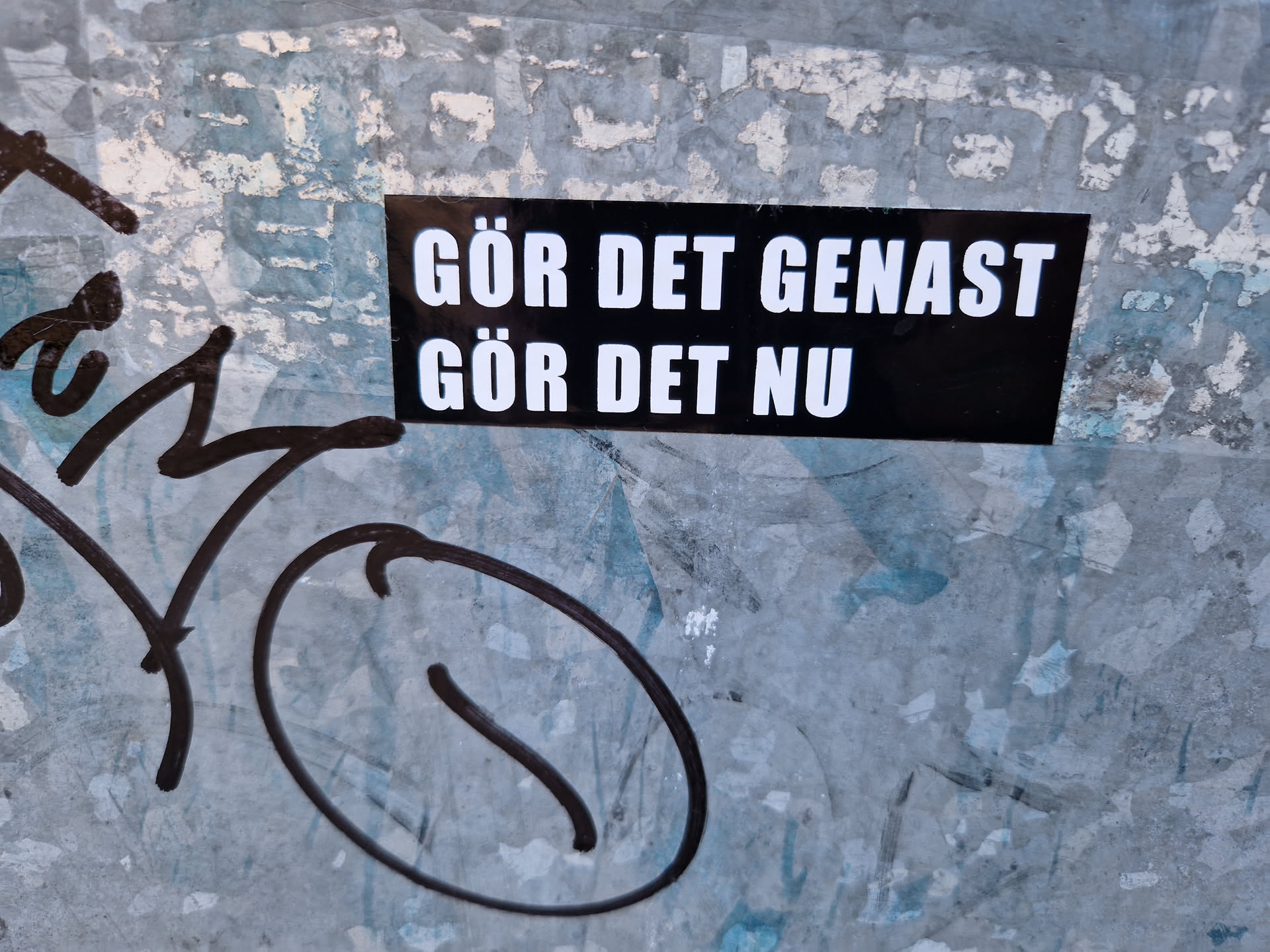 A black sticker with white text on a weathered surface. The text is in Swedish and translates to "Do it immediately, do it now."