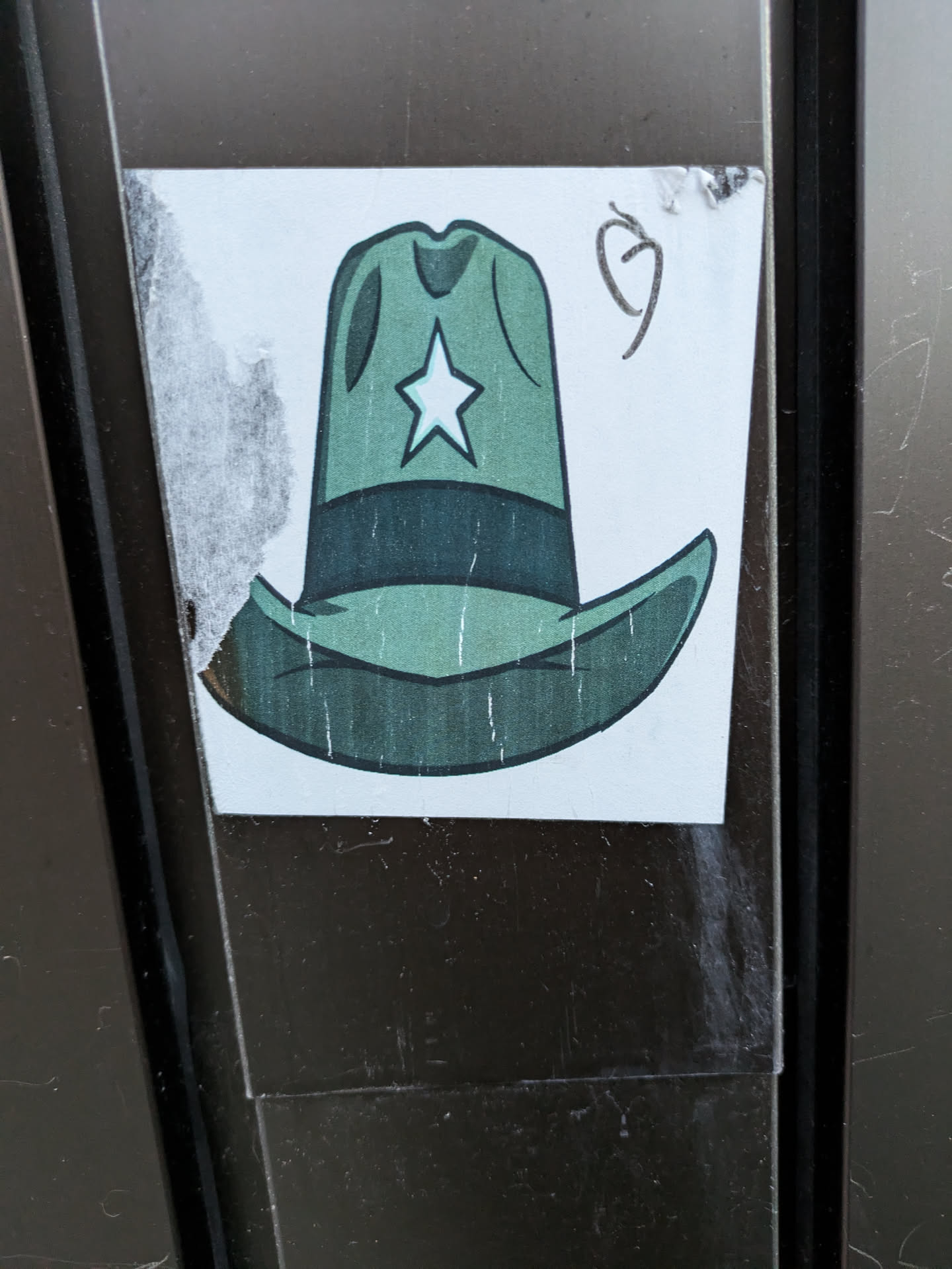 A sticker featuring a cartoon illustration of a green cowboy hat with a white star on the crown. The sticker appears to be hand-drawn or digitally created with a simple, slightly rustic style. It's affixed to a dark brown surface, showing some signs of wear and tear around the edges.