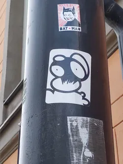 Close-up shot of a sticker featuring a stylized creature with large, round eyes and a simplistic design. The creature is predominantly black with white highlights, giving a striking contrast. It appears to be a unique character with no obvious reference to an existing character. The sticker is affixed to a dark-colored pole, along with other stickers.