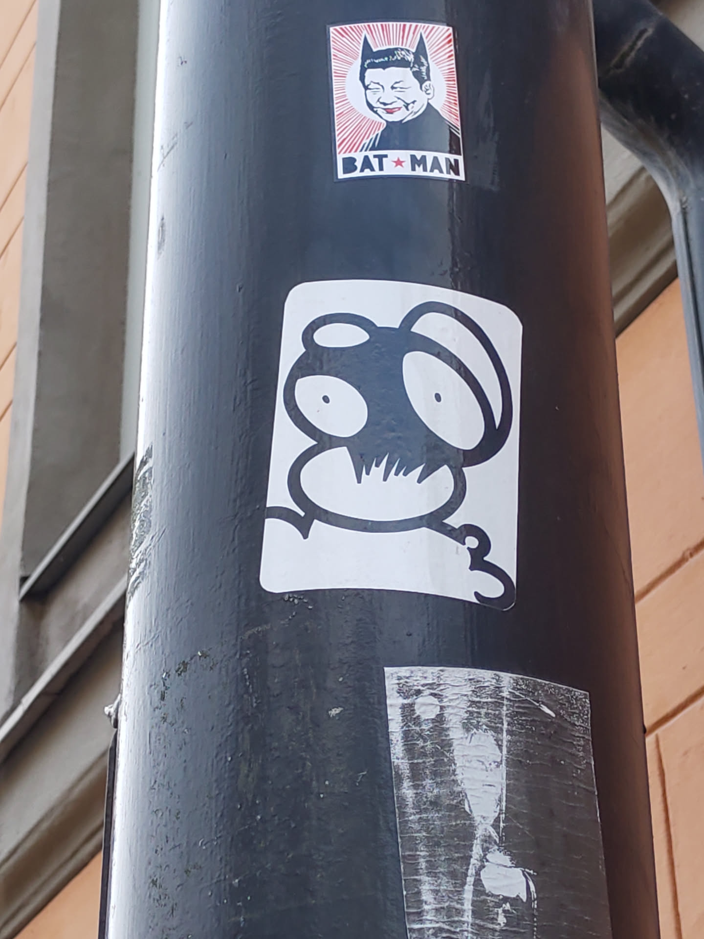 Close-up shot of a sticker featuring a stylized creature with large, round eyes and a simplistic design. The creature is predominantly black with white highlights, giving a striking contrast. It appears to be a unique character with no obvious reference to an existing character. The sticker is affixed to a dark-colored pole, along with other stickers.