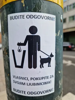 A sticker reminding dog owners to clean up after their pets.  It features a pictogram of a person picking up dog waste and placing it in a bin.  The text is in Croatian.