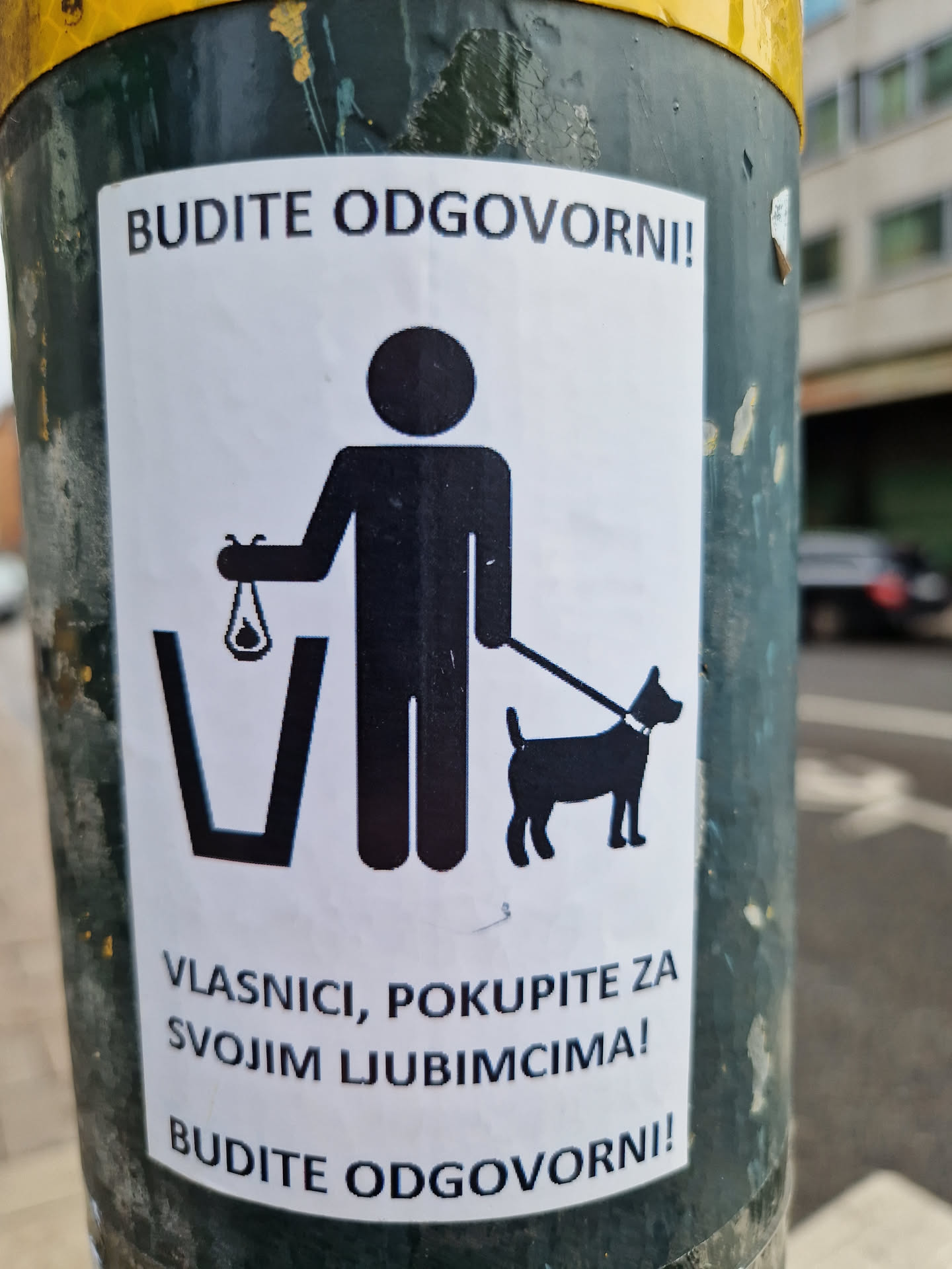 A sticker reminding dog owners to clean up after their pets.  It features a pictogram of a person picking up dog waste and placing it in a bin.  The text is in Croatian.