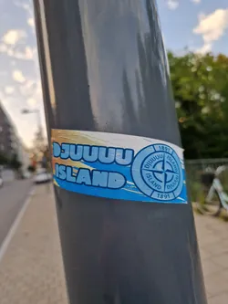 A blue and white sticker featuring the text 'DJUUUUU ISLAND' in a stylized font. A circular logo with a compass design and the text 'DJUUUUU ISLAND 1891' is also present. The sticker is affixed to a grey pole outdoors.