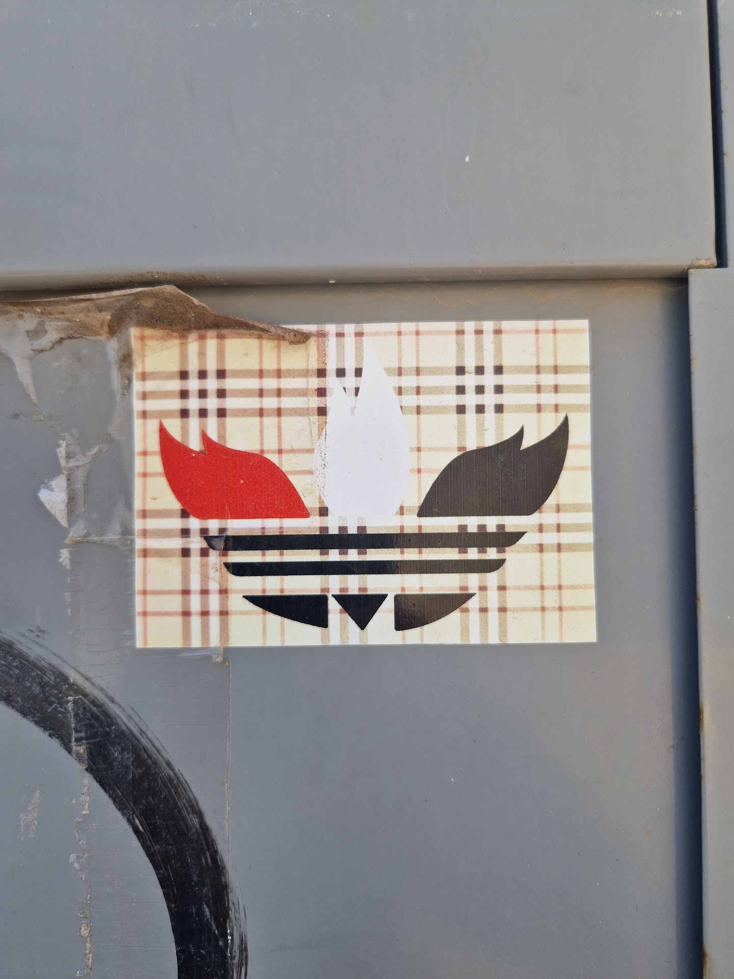 A sticker featuring a stylized Adidas logo overlaid on a Burberry-style plaid background. The Adidas logo is rendered in black, white, and red. The sticker is affixed to a gray surface.