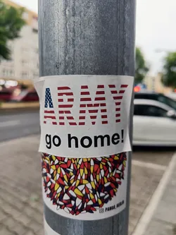 A sticker affixed to a pole displays the text "ARMY go home!" in red and white letters against a background resembling the American flag. Below this is an abstract design in red, yellow, white and blue.  A small artist's mark is visible at the bottom, possibly indicating a social media handle.